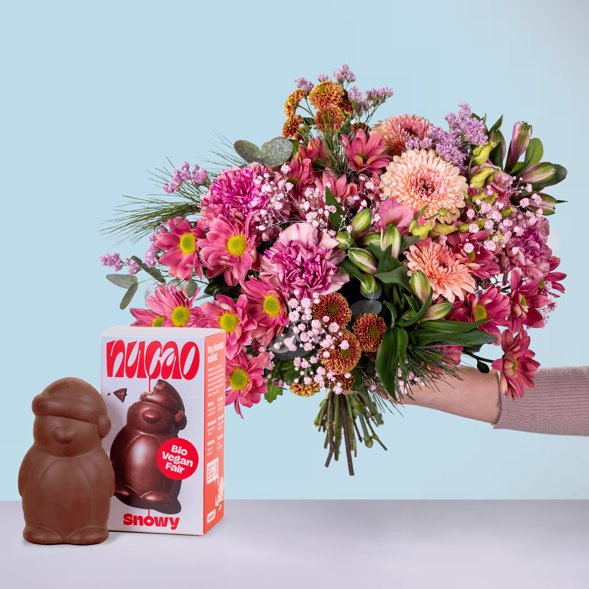 Ein leuchtender Strauß rosa, orangefarbener und violetter Blumen wird vor einem hellblauen Hintergrund vor einer Schachtel Yucao-Schokoladenbonbons gehalten. (Beschreibungstext KI-generiert)