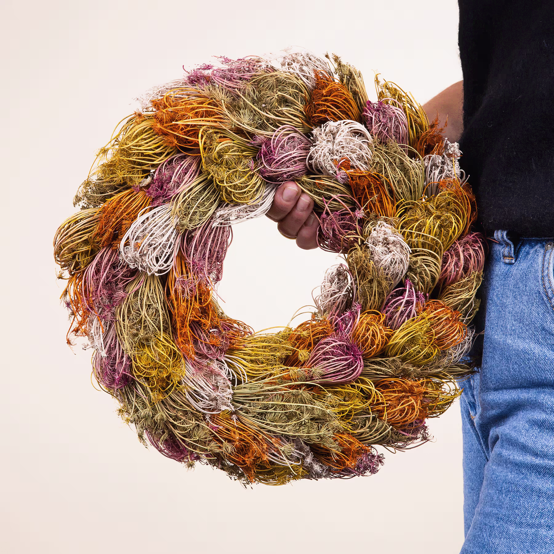 Ein farbenfroher, handgefertigter Kranz aus verschiedenen bunten Trockenblumen und Blättern, gehalten von einer Person in Jeans im Vordergrund. (Beschreibungstext KI-generiert)