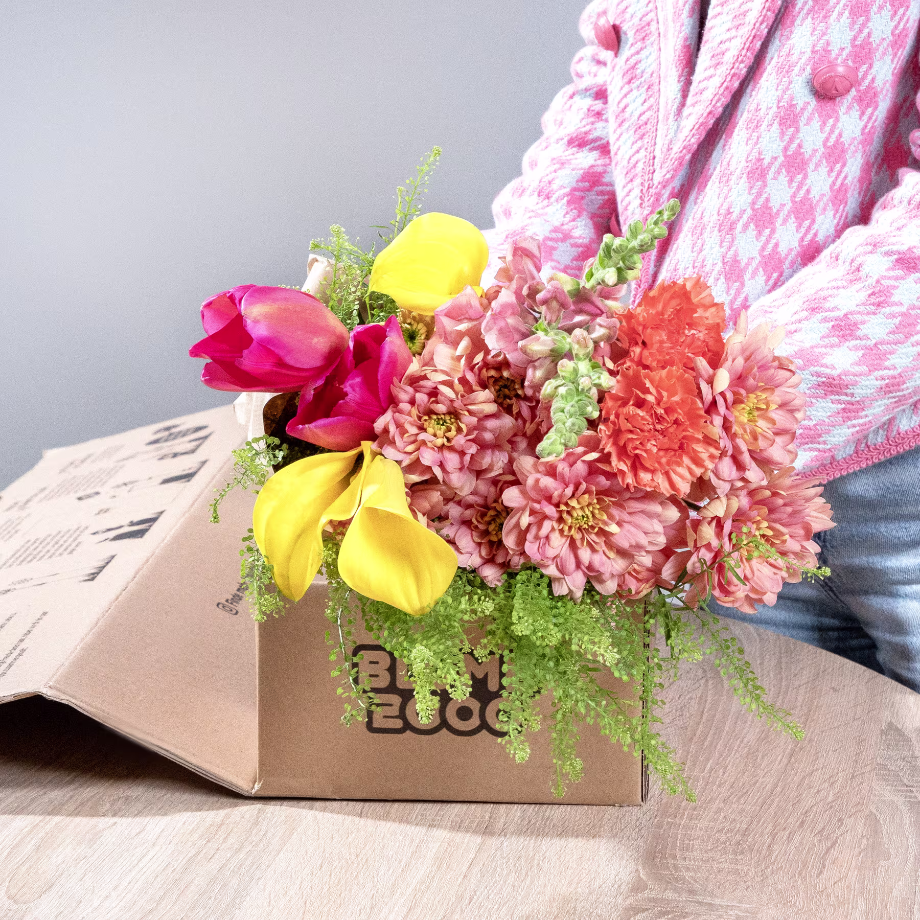 Ein leuchtender Strauß bunter Blumen, darunter rote Tulpen, gelbe Narzissen und rosa Chrysanthemen, steht in einem Karton auf einer Holzoberfläche, im Hintergrund ist ein rosa-weiß karierter Stoff zu sehen. (Beschreibungstext KI-generiert)