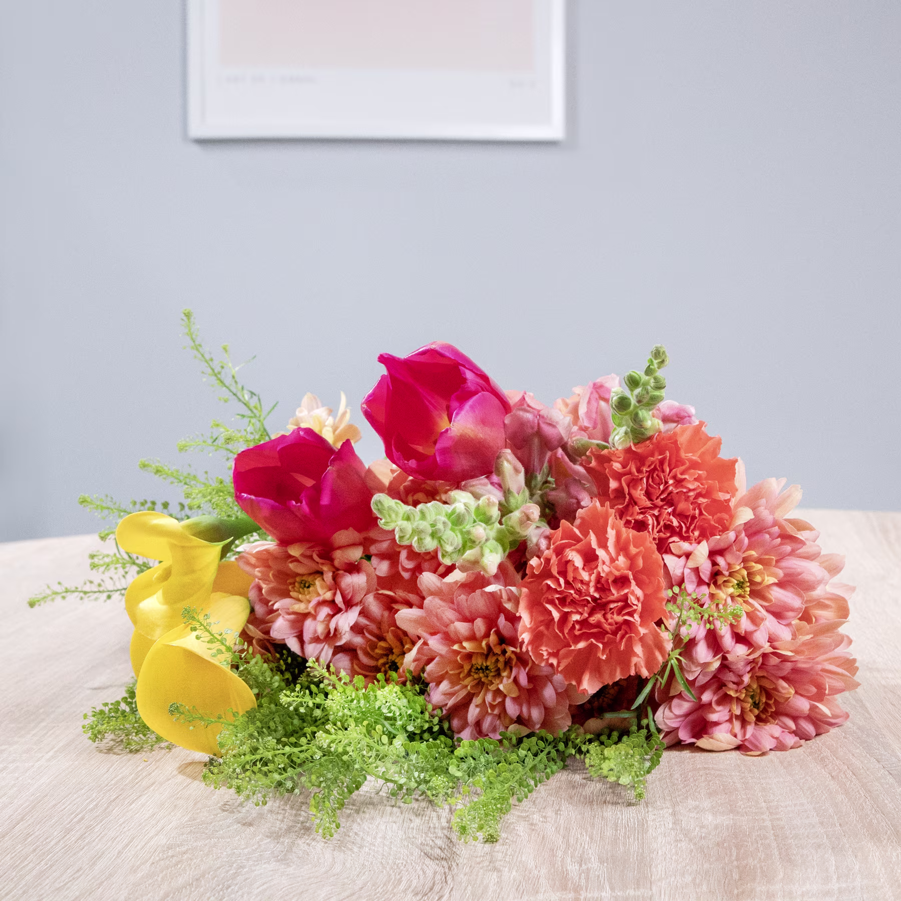 Ein farbenfroher Strauß roter Rosen, rosa Nelken und grünem Laub steht auf einem Holztisch vor einer schlichten weißen Wand mit einem gerahmten Kunstwerk. (Beschreibungstext KI-generiert)