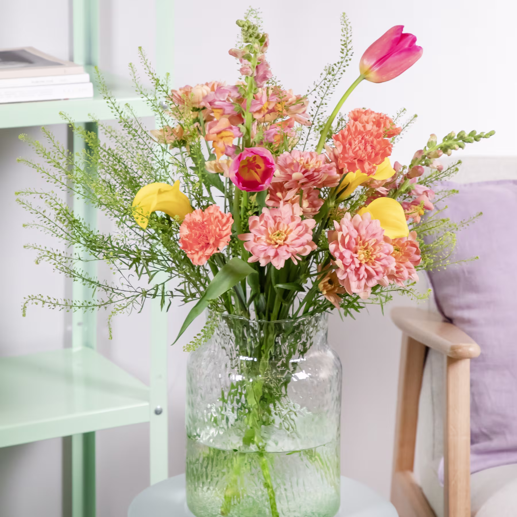 Ein leuchtender Strauß bunter Blumen, darunter Tulpen, Gänseblümchen und Nelken, wird in einer Glasvase vor dem Hintergrund eines Holzstuhls und eines grünen Regals präsentiert. (Beschreibungstext KI-generiert)