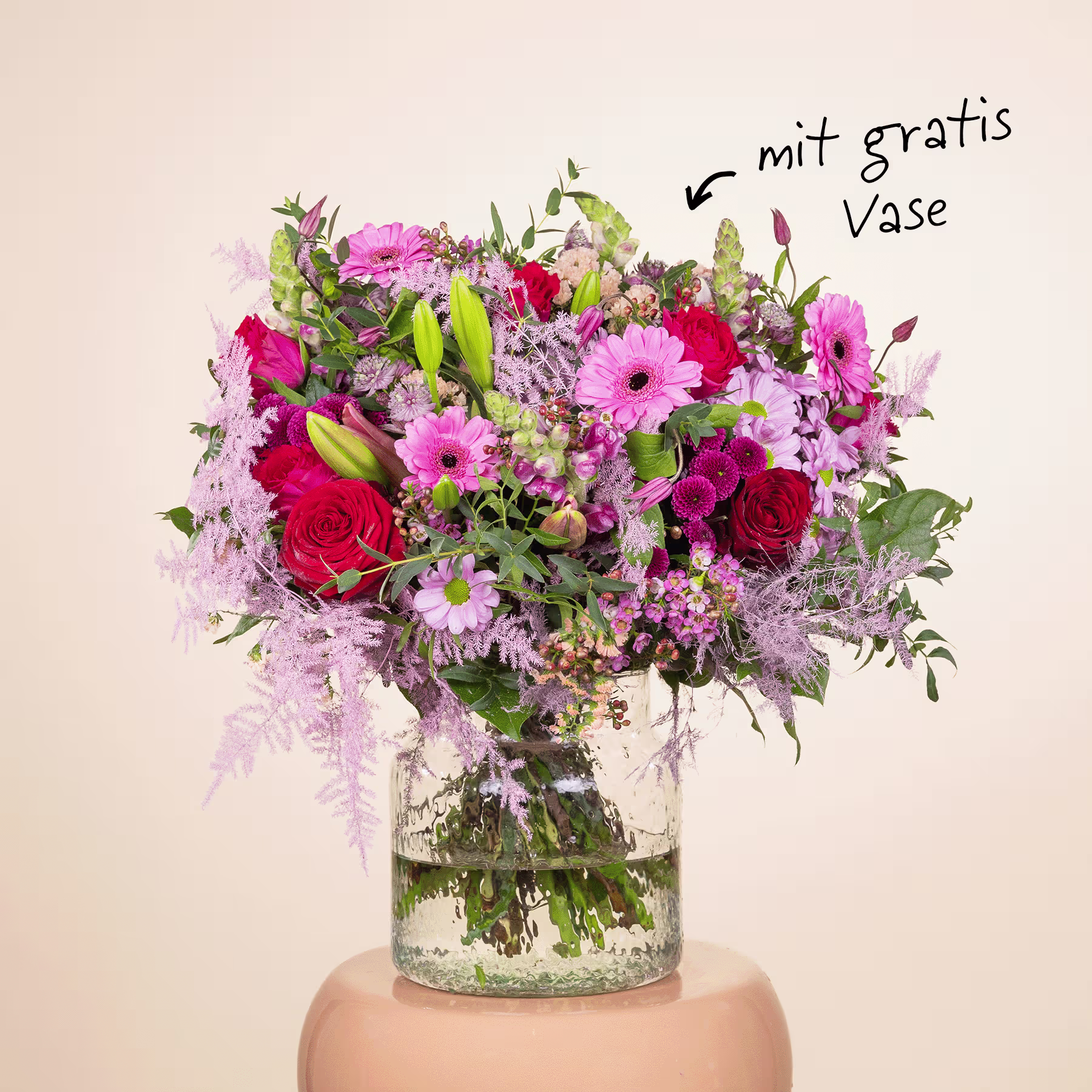 Ein lebendiger Strauß verschiedener bunter Blumen, darunter rosa Gerbera, rote Rosen sowie violette und weiße Blumen, wird in einer Glasvase vor einem schlichten beigen Hintergrund präsentiert. (Beschreibungstext KI-generiert)