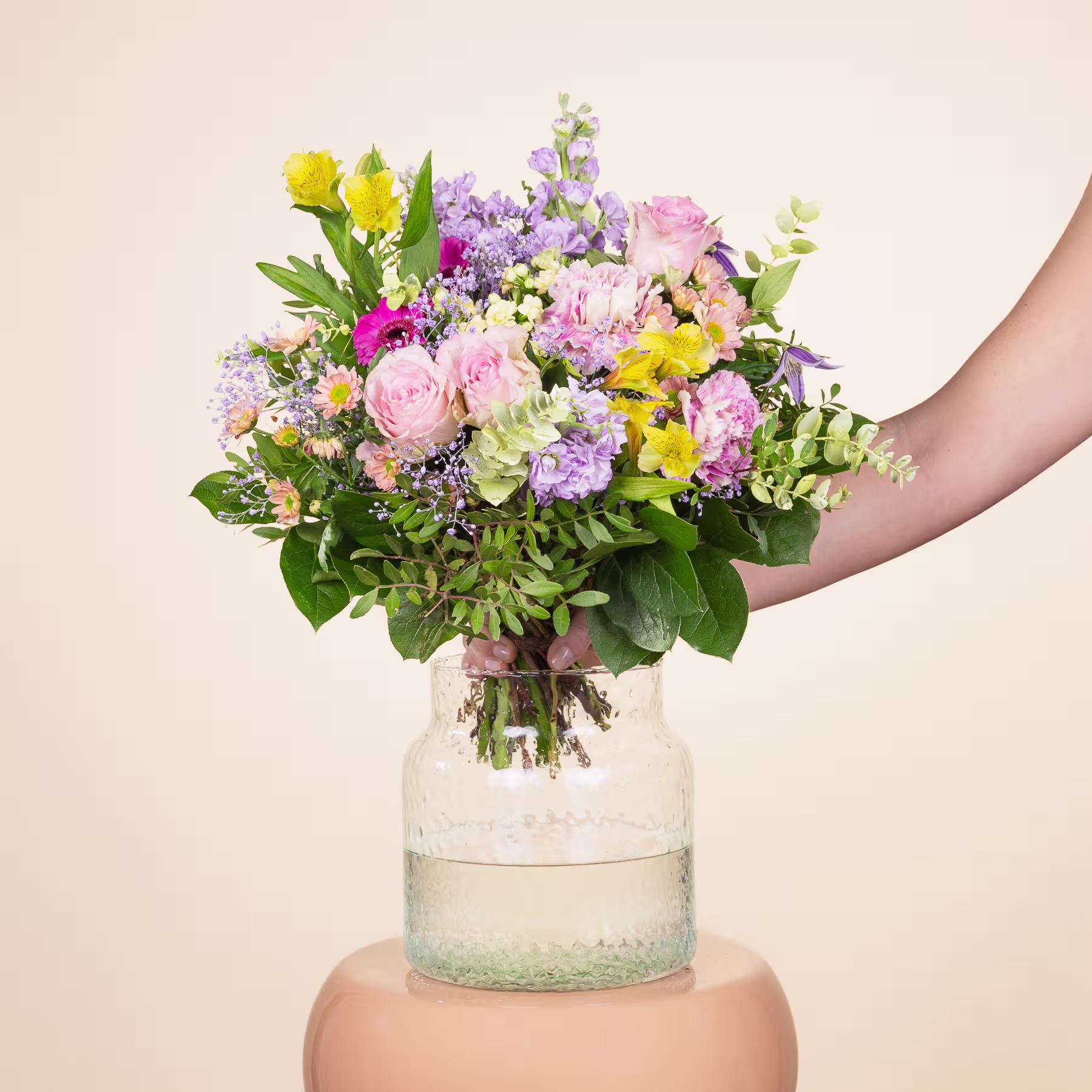 Ein lebendiger Strauß verschiedener bunter Blumen, darunter Rosen, Nelken und andere Blüten, steht in einer Glasvase vor einem schlichten, hellen Hintergrund. (Beschreibungstext KI-generiert)