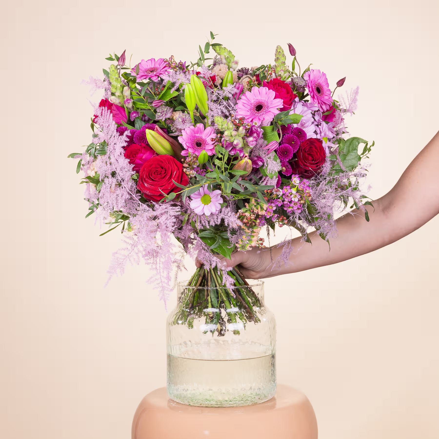 Ein farbenfroher Strauß rosa und roter Blumen, darunter Rosen und Gerbera, steht in einer Glasvase vor einem schlichten beigen Hintergrund. (Beschreibungstext KI-generiert)