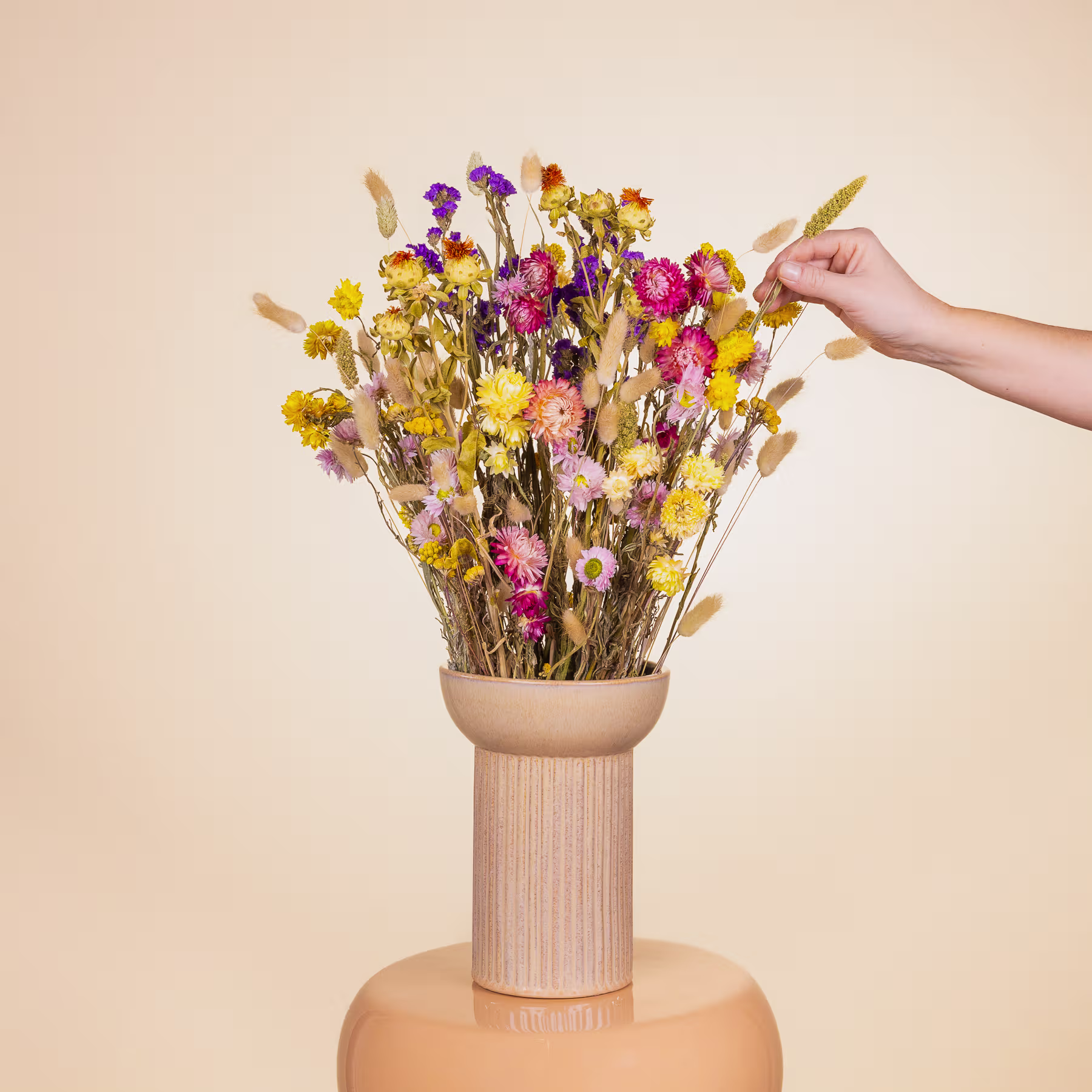 Eine Hand hält vor einem schlichten beigen Hintergrund eine Vase mit einem farbenfrohen Strauß getrockneter Blumen in Gelb-, Rosa- und Lilatönen. (Beschreibungstext KI-generiert)