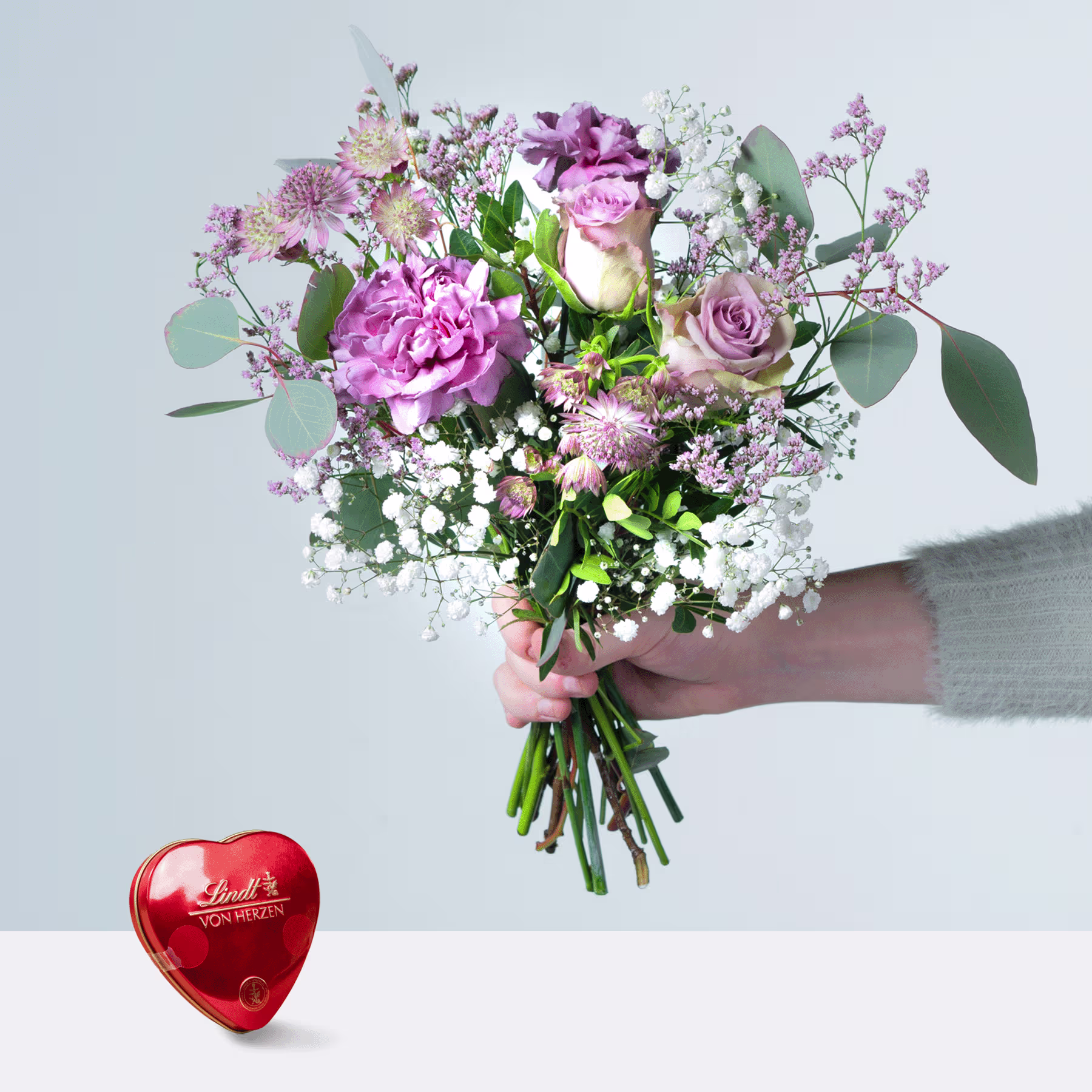 Eine Person hält einen farbenfrohen Strauß rosa und violetter Blumen, darunter Rosen und Pfingstrosen, in den Händen, im Vordergrund steht eine rote, herzförmige Pralinenschachtel. (Beschreibungstext KI-generiert)