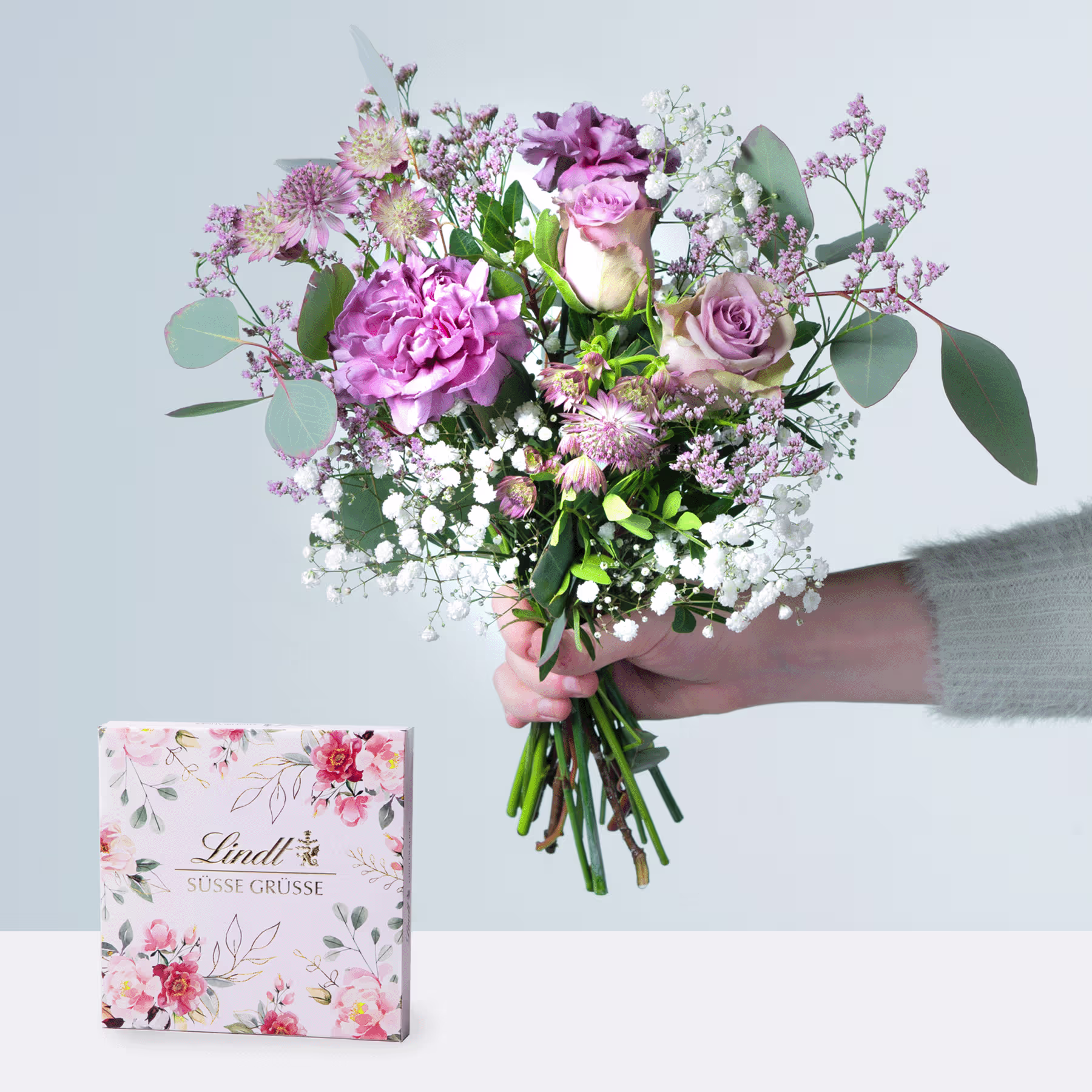 Eine Person hält einen farbenfrohen Strauß rosa und violetter Blumen, darunter Rosen und Pfingstrosen, in den Händen, begleitet von einer Schachtel Lindt-Pralinen auf einer weißen Oberfläche. (Beschreibungstext KI-generiert)