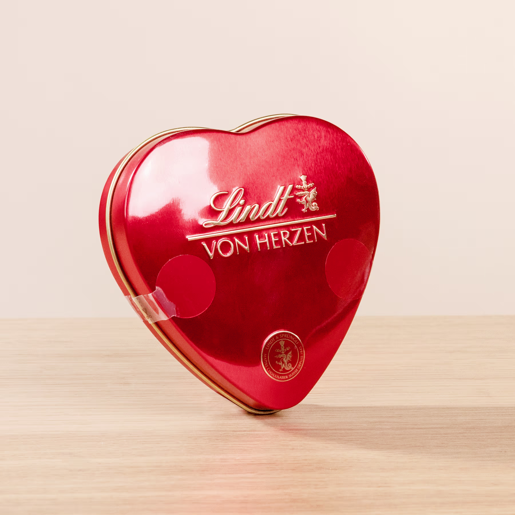Eine rote herzförmige Dose mit Lindt-Pralinen „Von Herzen“ steht auf einer Holzoberfläche. (Beschreibungstext KI-generiert)