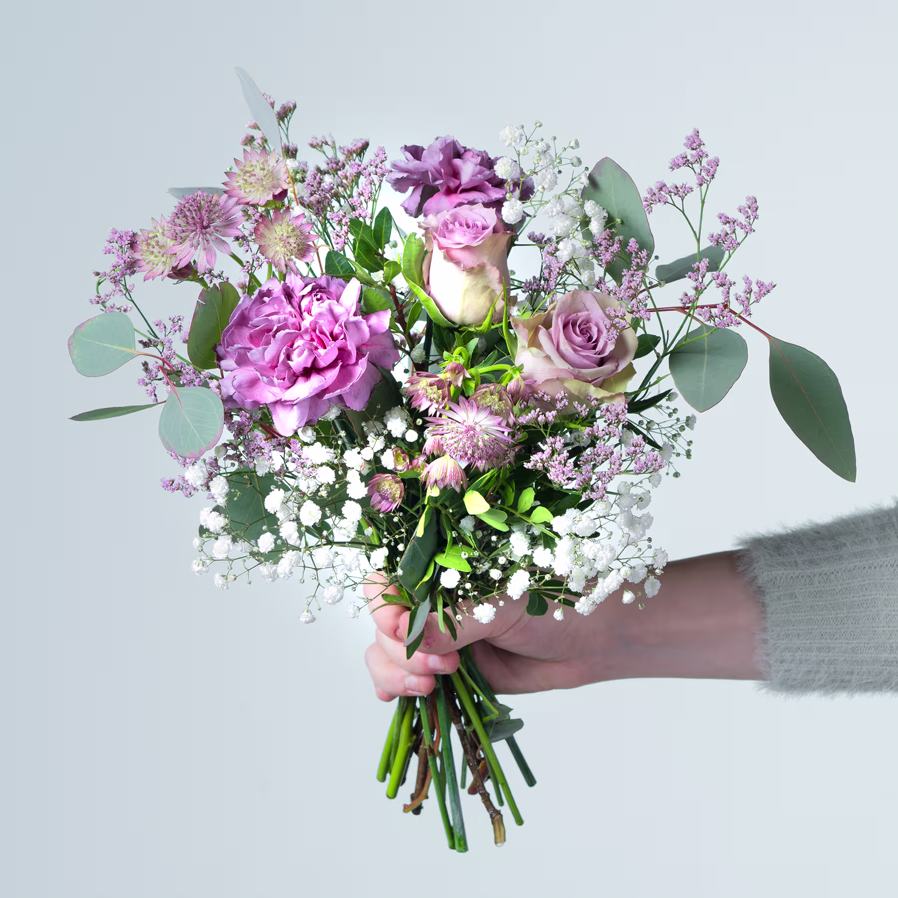 Ein leuchtender Strauß rosa und violetter Blumen, darunter Pfingstrosen, Rosen und andere zarte Blüten, wird vor einem schlichten, hellen Hintergrund in den Händen einer Person gehalten. (Beschreibungstext KI-generiert)
