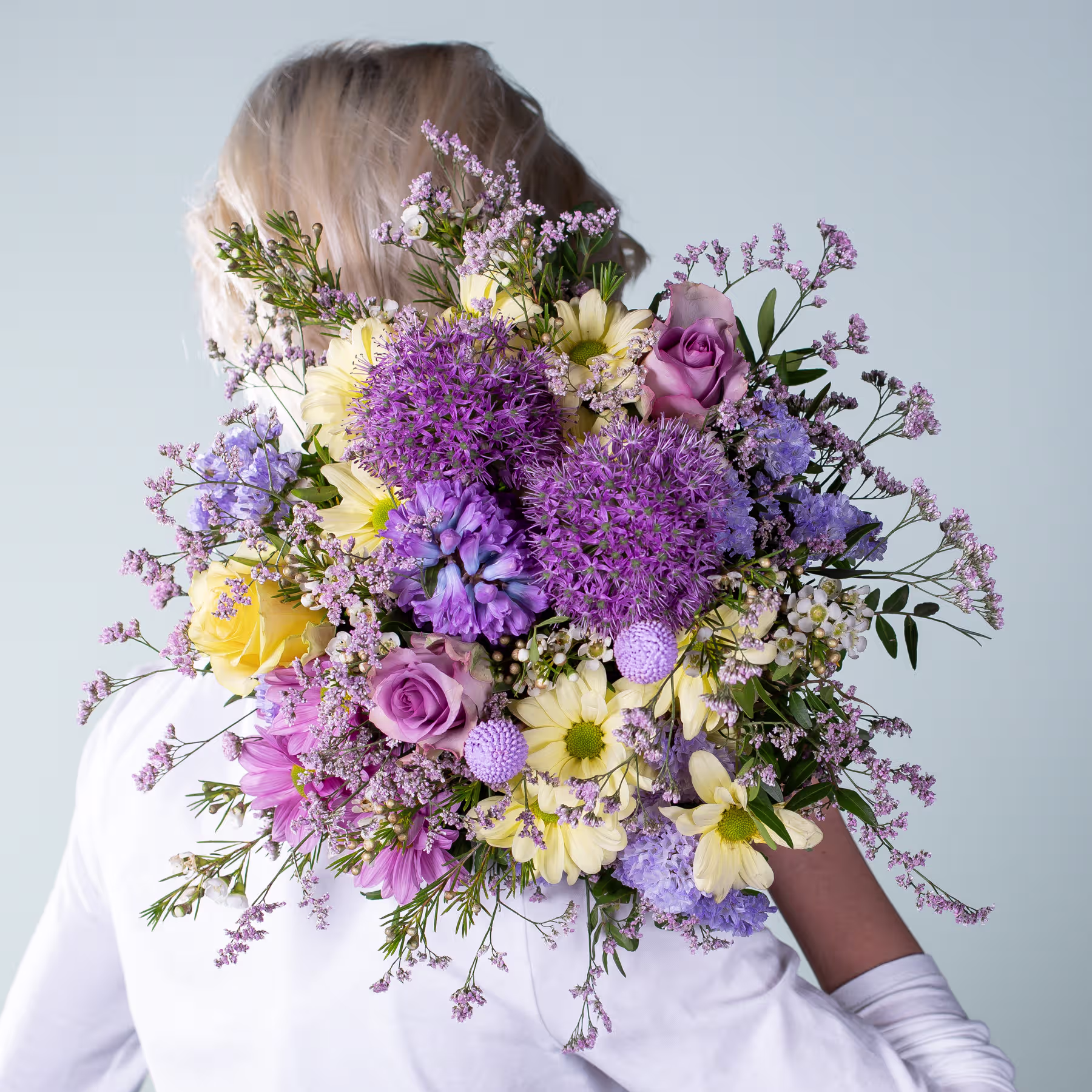 Ein leuchtender Strauß bunter Blumen, darunter violette Chrysanthemen, gelbe Narzissen und rosa Rosen, gehalten von einer Person in einem weißen Hemd vor einem schlichten Hintergrund. (Beschreibungstext KI-generiert)