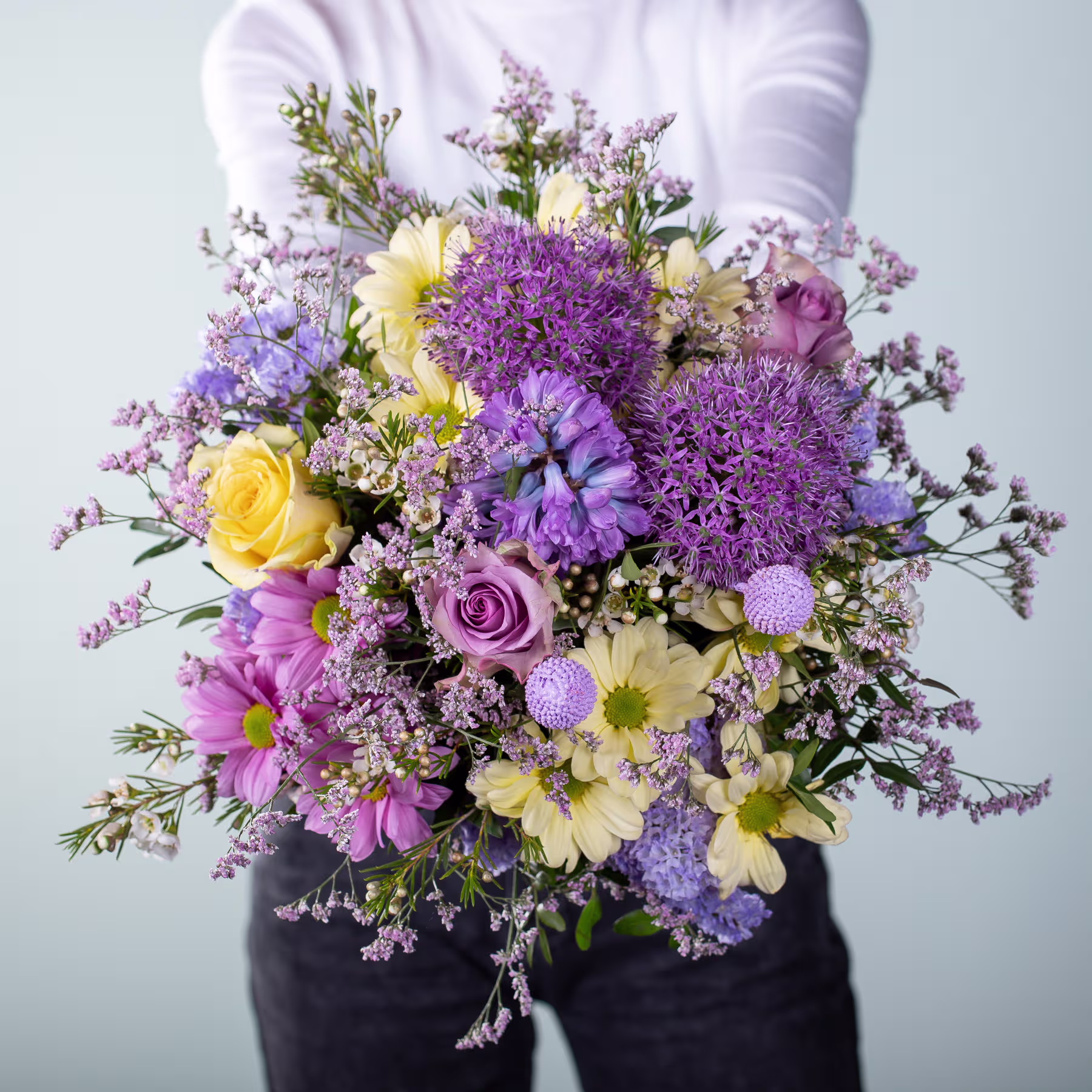 Ein leuchtender Strauß bunter Blumen, darunter lila Allium, gelbe Narzissen und rosa Rosen, wird vor einem schlichten Hintergrund in den Händen einer Person gehalten. (Beschreibungstext KI-generiert)