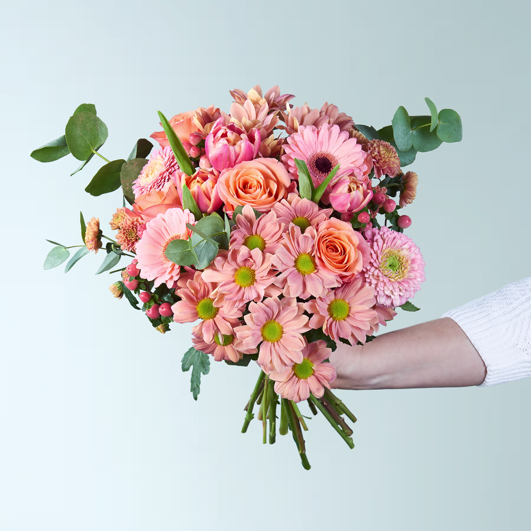 Ein leuchtender Strauß rosa und orangefarbener Blumen, darunter Rosen und Gänseblümchen, wird vor einem hellblauen Hintergrund in den Händen einer Person gehalten. (Beschreibungstext KI-generiert)