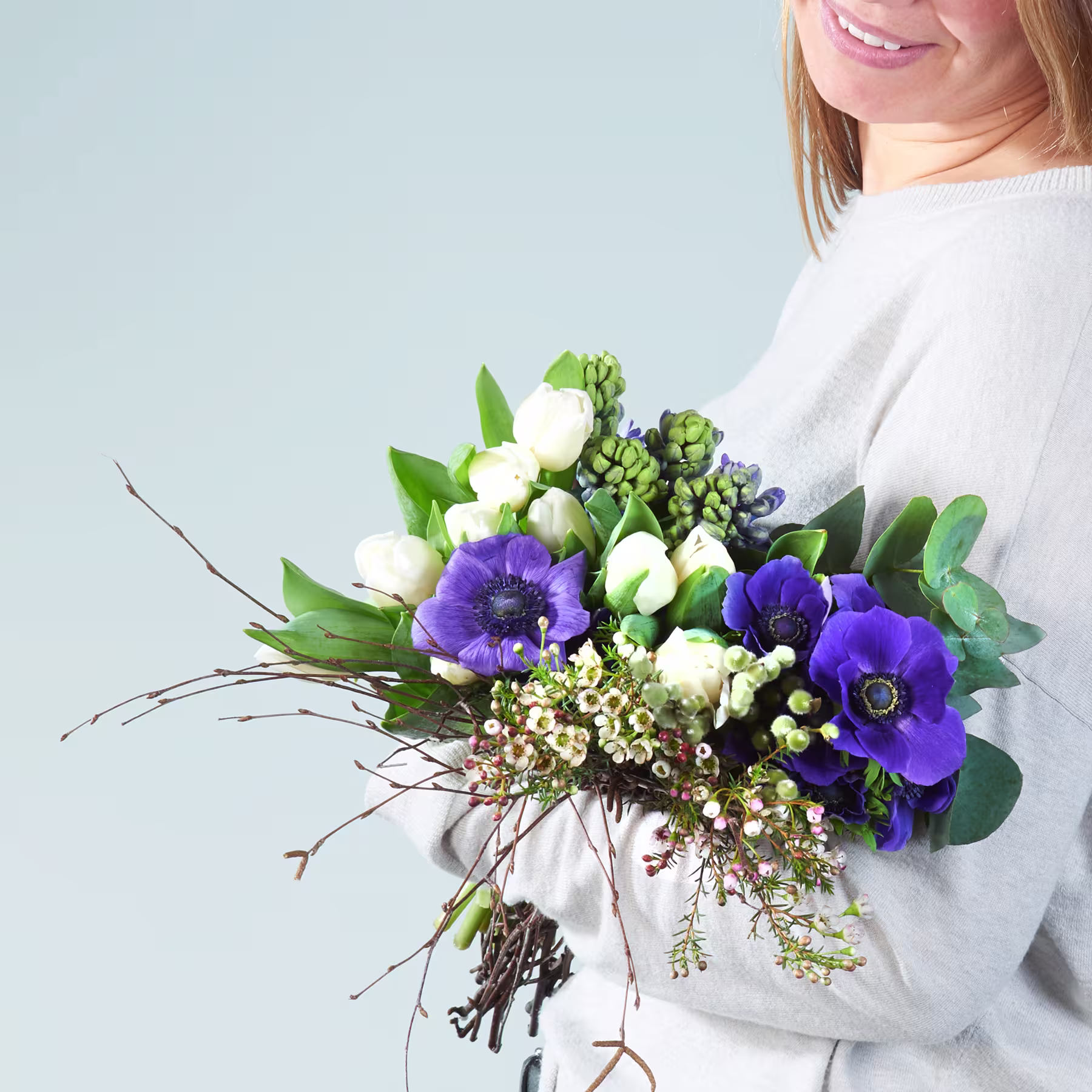 Eine Frau in einem weißen Pullover hält vor einem schlichten hellblauen Hintergrund einen farbenfrohen Strauß weißer und violetter Blumen, darunter Anemonen und andere Grünpflanzen. (Beschreibungstext KI-generiert)