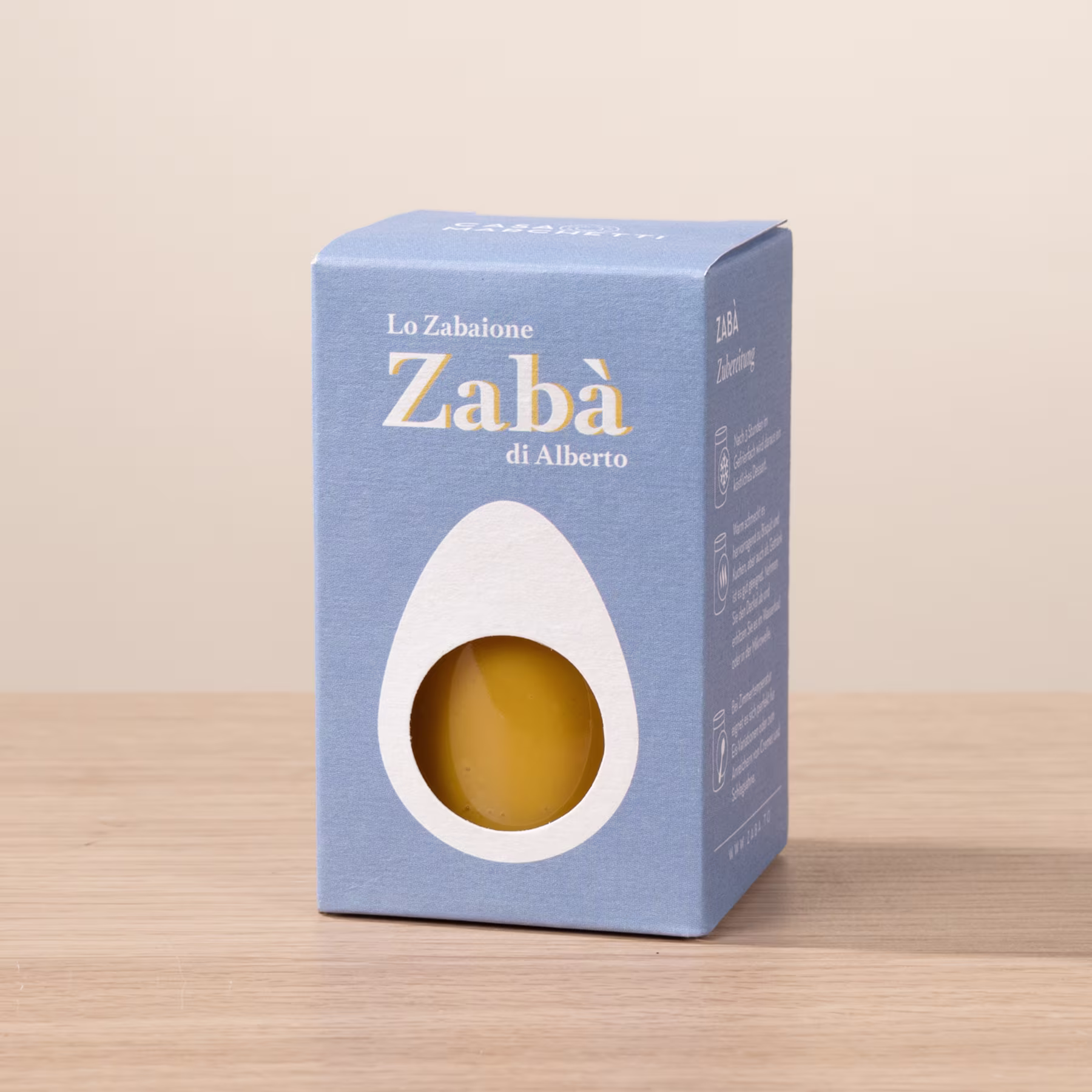 Ein blauer Karton mit dem Text „Zaba di Alberto“ und einem gelben Gegenstand darin, der auf einer Holzoberfläche ruht. (Beschreibungstext KI-generiert)