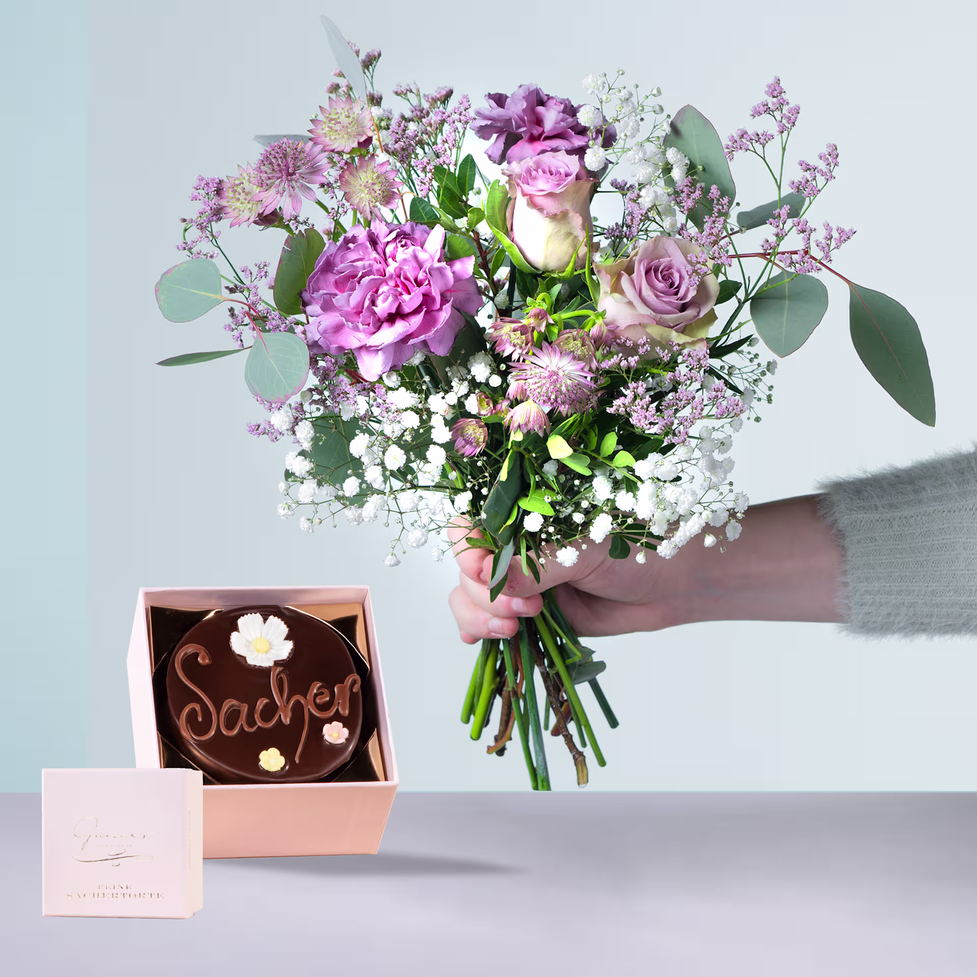 Eine Person hält einen farbenfrohen Strauß rosa und weißer Blumen, darunter Rosen und Pfingstrosen, in den Händen, zusammen mit einer kleinen Pralinenschachtel mit der Aufschrift „Sasher“. (Beschreibungstext KI-generiert)