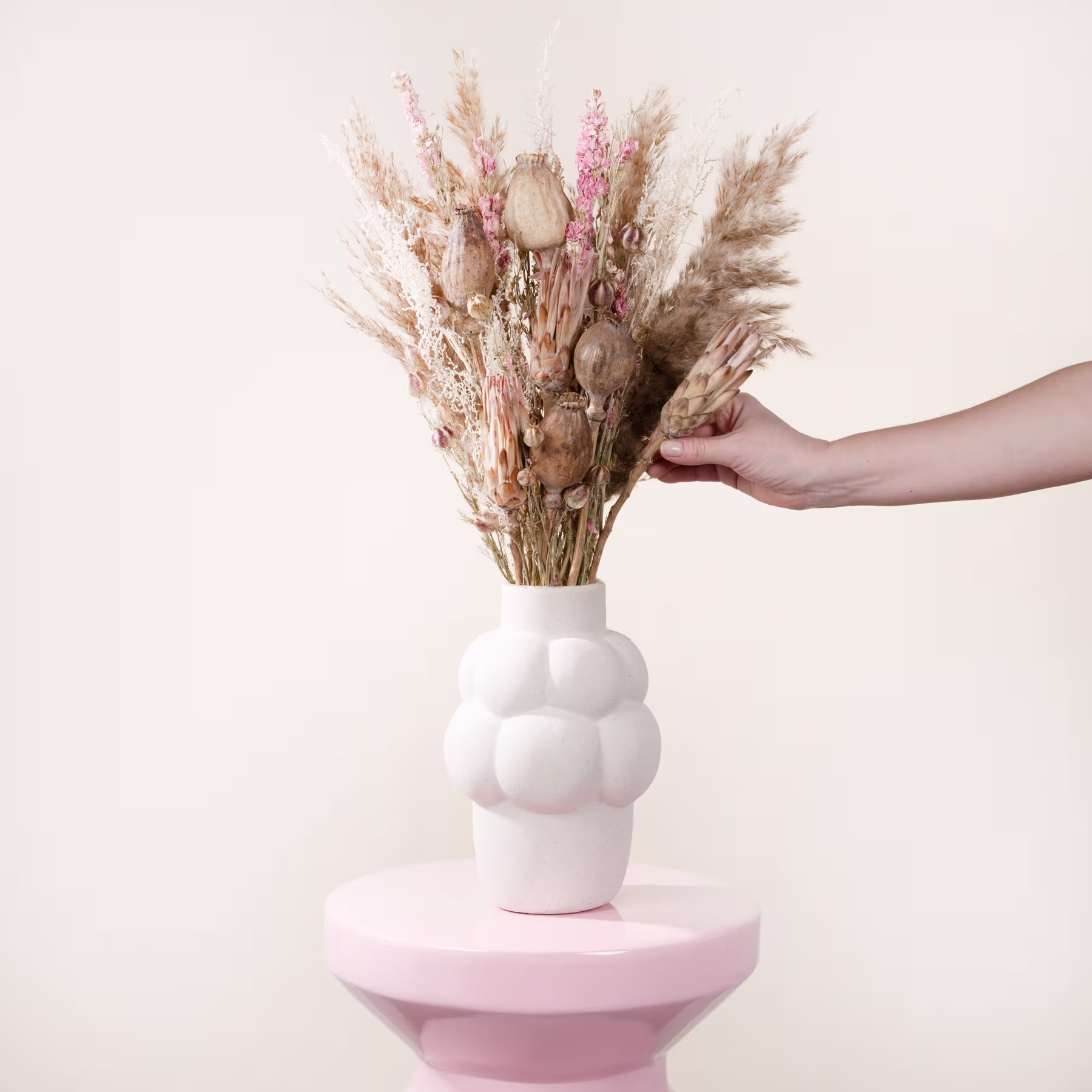 Eine Hand hält einen Strauß getrockneter Blumen in einer weißen Vase vor einem schlichten, hellen Hintergrund. (Beschreibungstext KI-generiert)