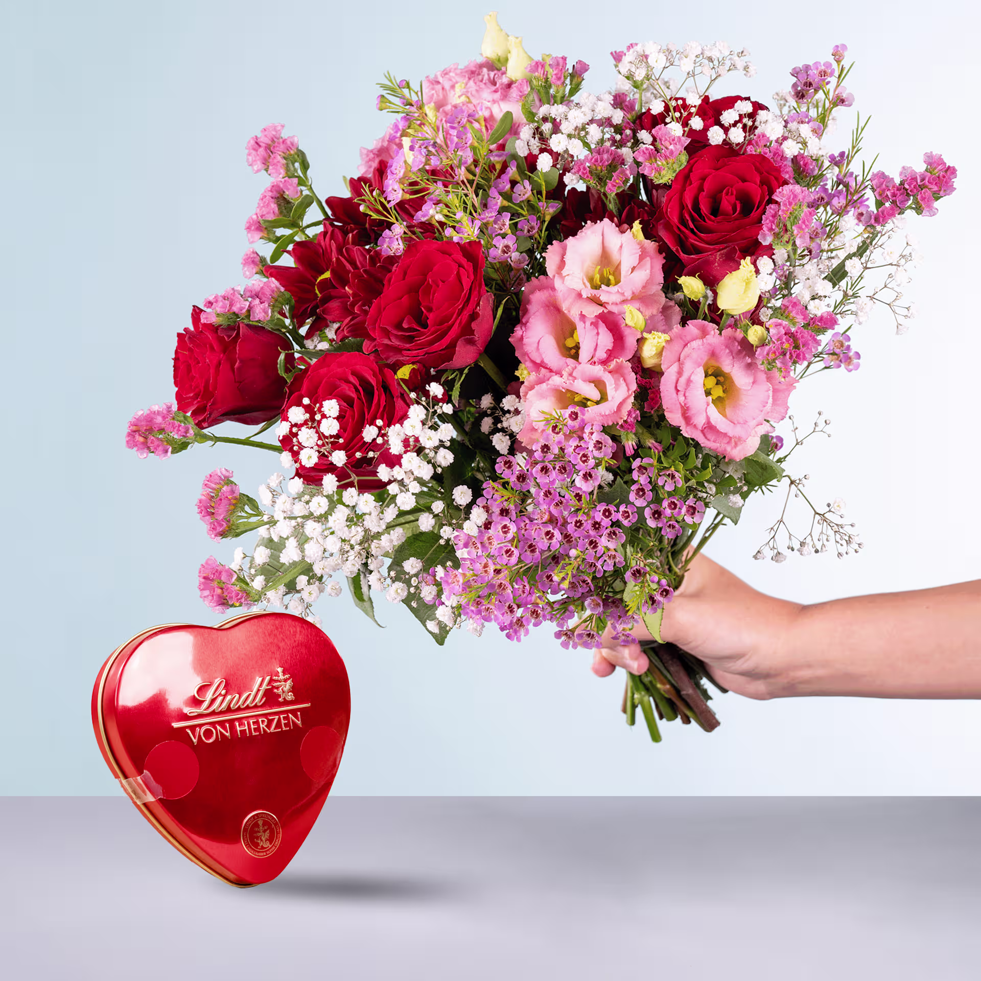 Eine Person hält einen leuchtenden Strauß roter Rosen, rosa Nelken und anderer bunter Blumen in den Händen, im Vordergrund ist vor einem schlichten Hintergrund eine herzförmige Lindt-Schokoladenschachtel zu sehen. (Beschreibungstext KI-generiert)