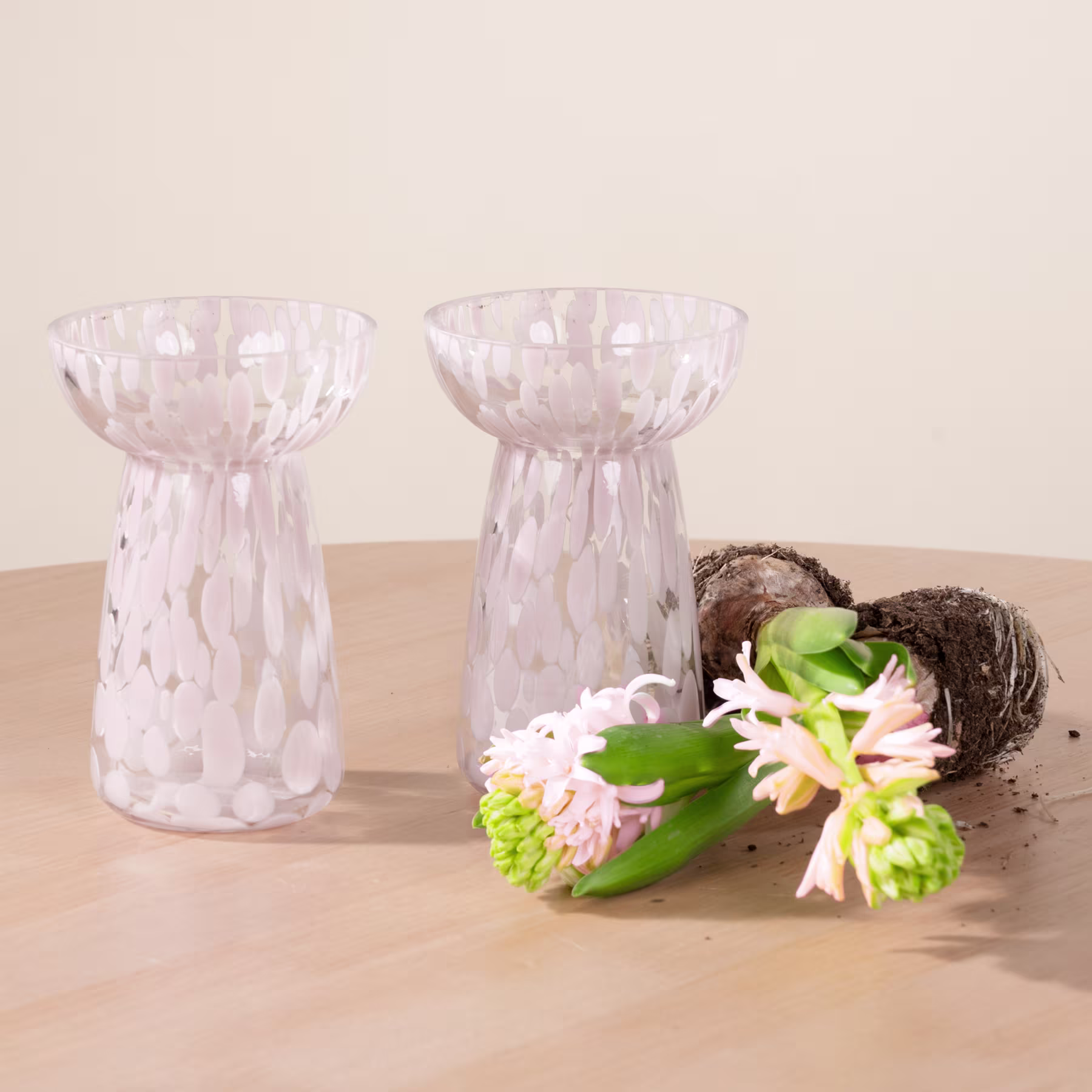Zwei klare Glasvasen mit strukturiertem Muster stehen auf einer Holzoberfläche, begleitet von einer kleinen nestartigen Struktur und einem Strauß leuchtend grüner und weißer Blumen. (Beschreibungstext KI-generiert)