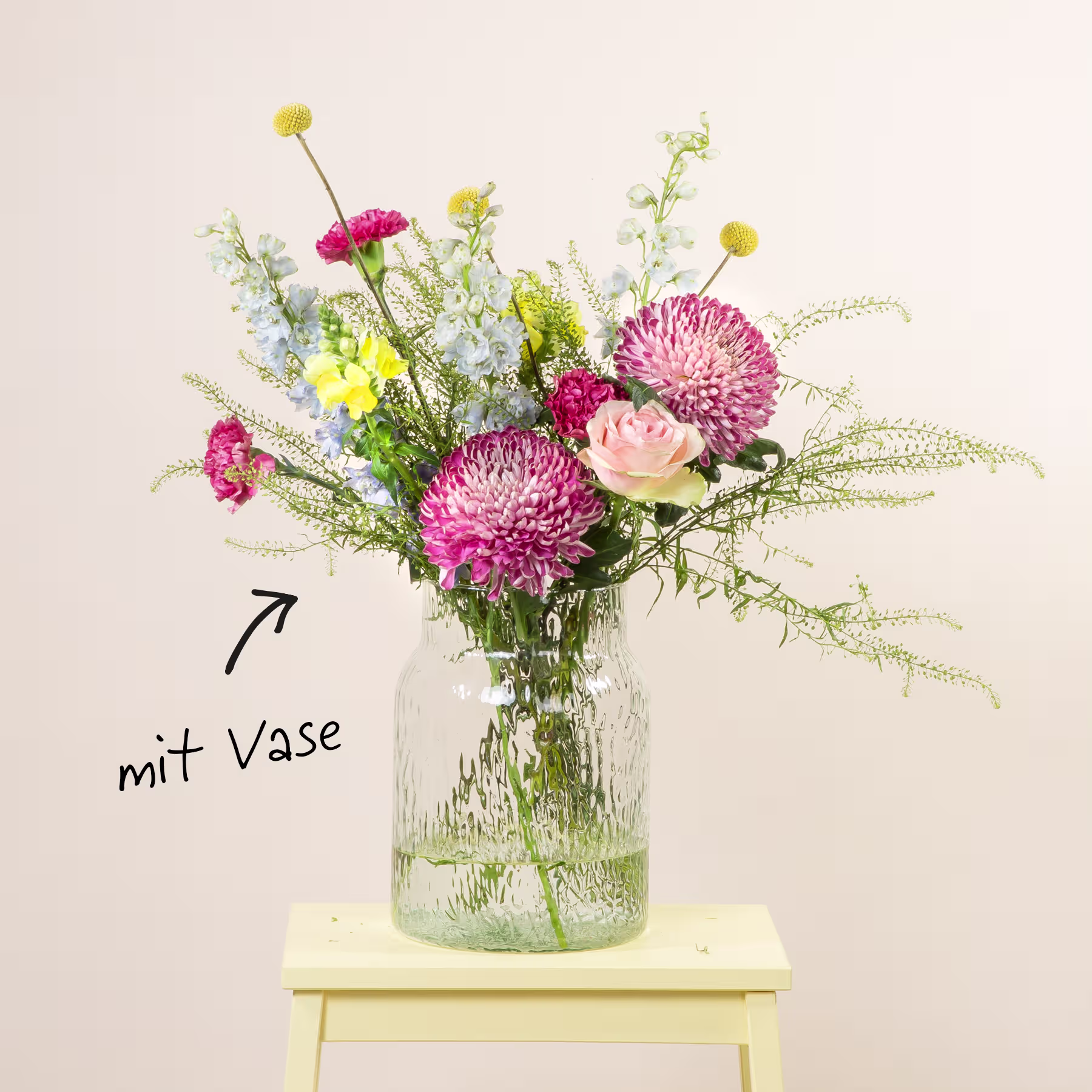 Ein Strauß leuchtend rosa und gelber Blumen, darunter Chrysanthemen und Rosen, wird in einer Glasvase auf einem gelben Holzhocker vor einem schlichten weißen Hintergrund präsentiert. (Beschreibungstext KI-generiert)