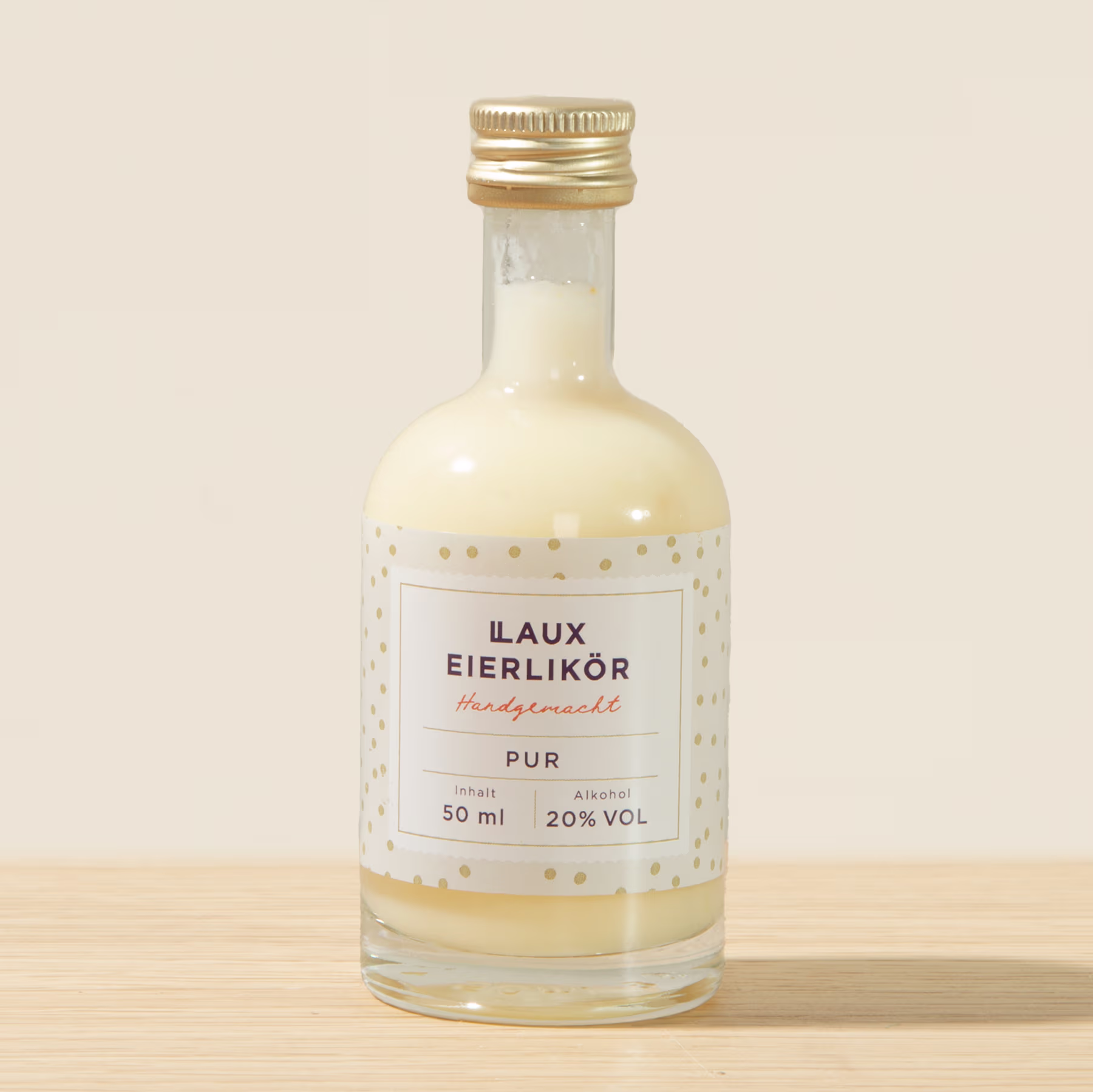 Eine kleine Glasflasche mit goldfarbenem Verschluss, die eine klare Flüssigkeit mit der Aufschrift „Laux Eierlikor Pur“ enthält, steht auf einer Holzoberfläche vor einem hellen Hintergrund. (Beschreibungstext KI-generiert)