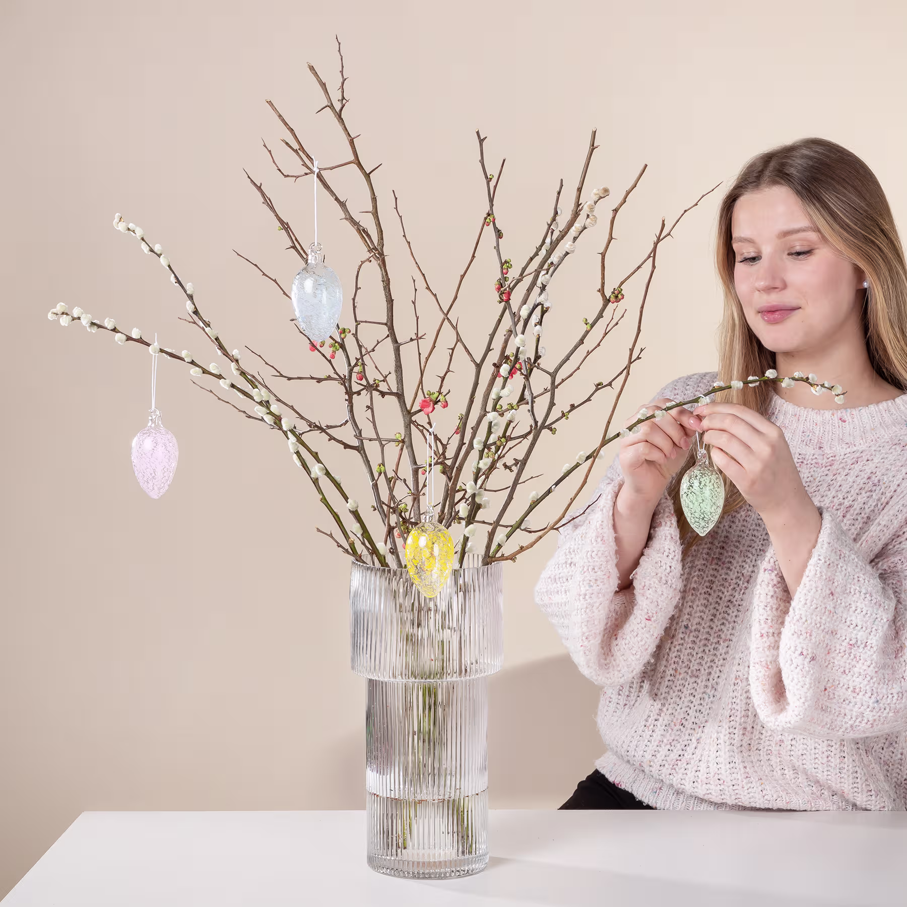 Eine junge Frau in einem kuscheligen Pullover dekoriert vor einem neutralen Hintergrund eine Vase mit kahlen Zweigen, die mit bunten Ostereiern geschmückt sind. (Beschreibungstext KI-generiert)