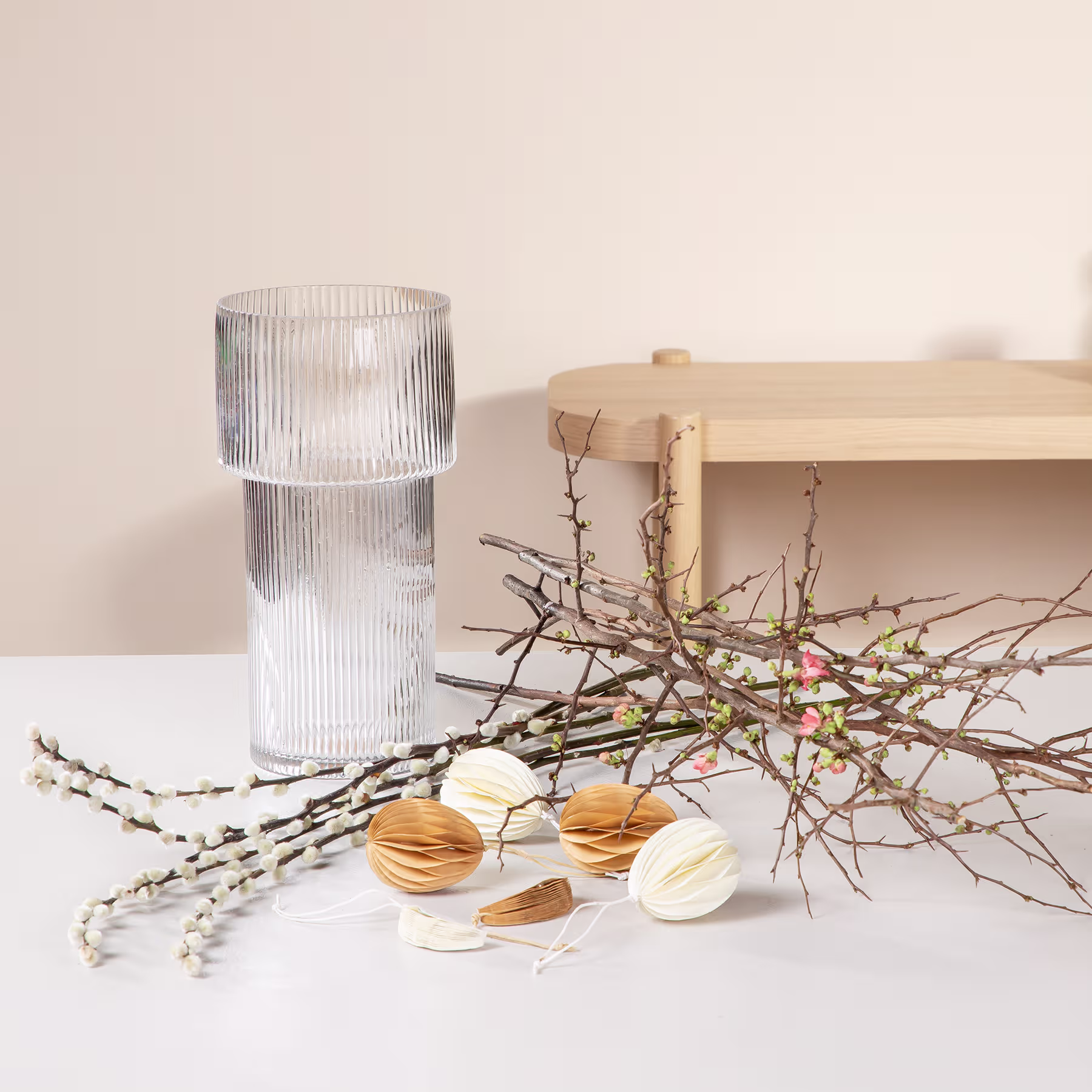 Auf einem Holzregal steht eine Glasvase mit Rippenmuster, daneben ein Zweig mit Frühlingsblüten und mehrere Macarons, die auf der Oberfläche angeordnet sind. (Beschreibungstext KI-generiert)