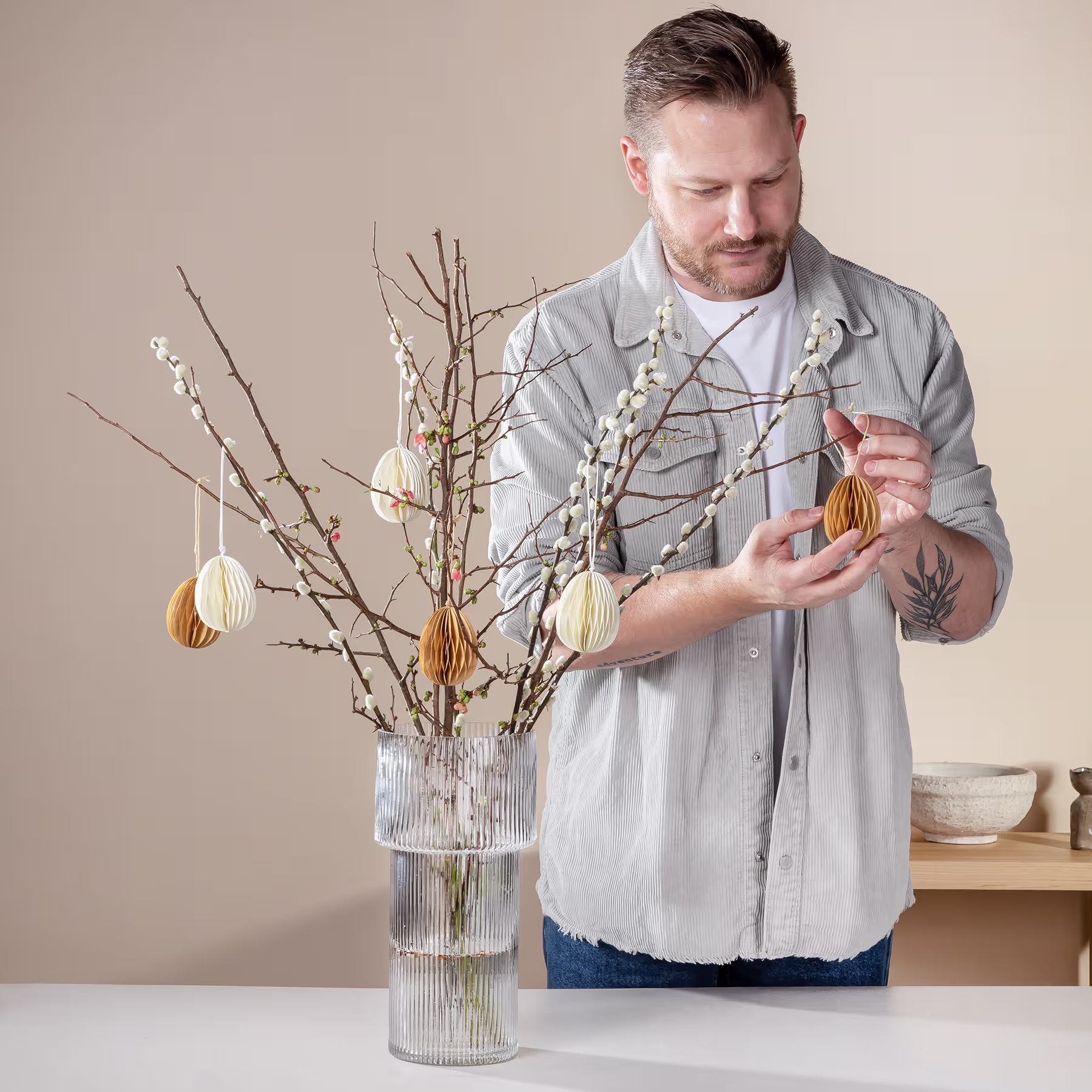 Ein Mann mit Bart und Tattoos arrangiert dekorative Zweige mit Ostereiern in einer Vase auf einem Tisch vor einem schlichten beigen Hintergrund. (Beschreibungstext KI-generiert)