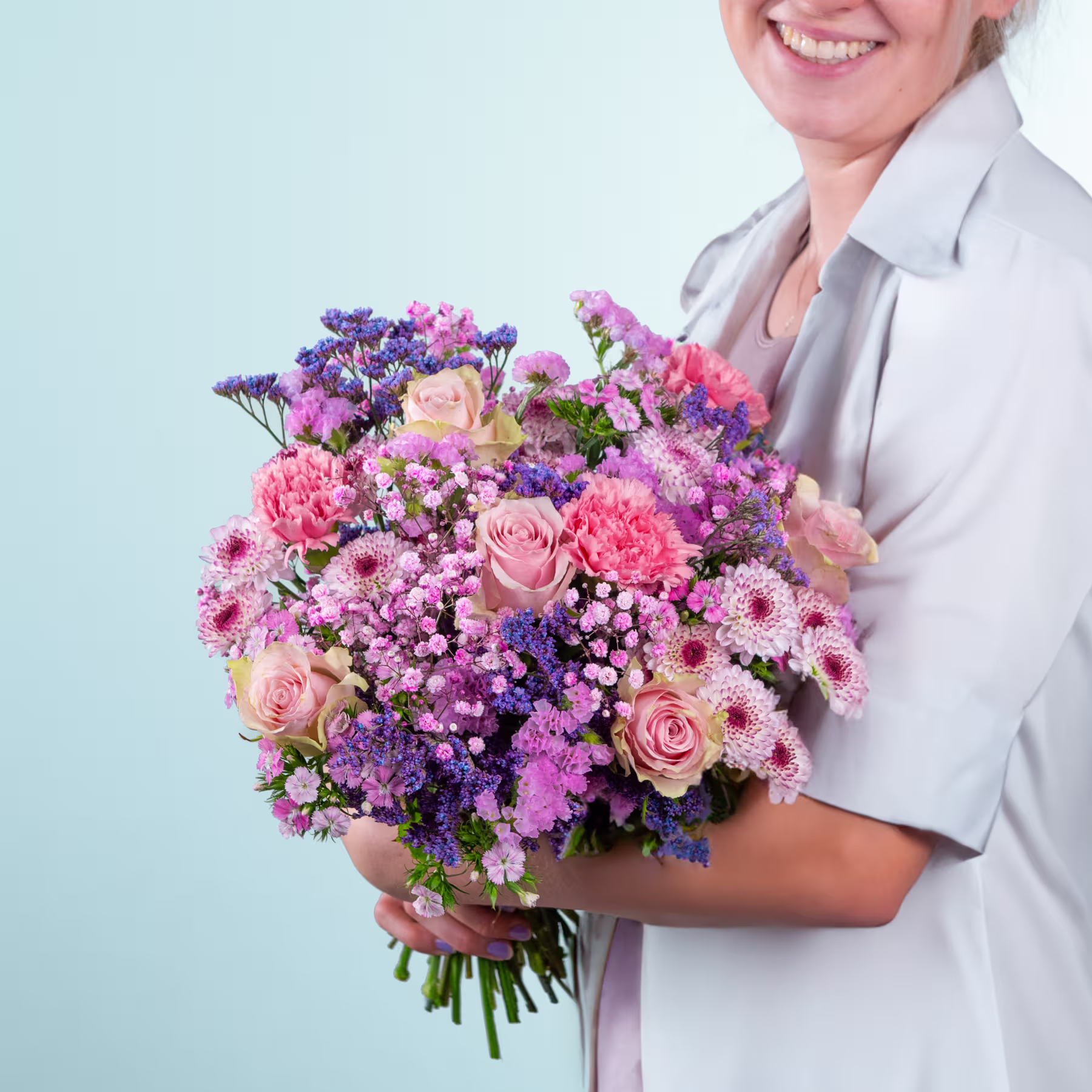 Eine lächelnde Frau in einer grauen Jacke hält vor einem hellblauen Hintergrund einen leuchtend bunten Blumenstrauß, darunter rosa Rosen, violette Schwertlilien und verschiedene andere Blüten. (Beschreibungstext KI-generiert)