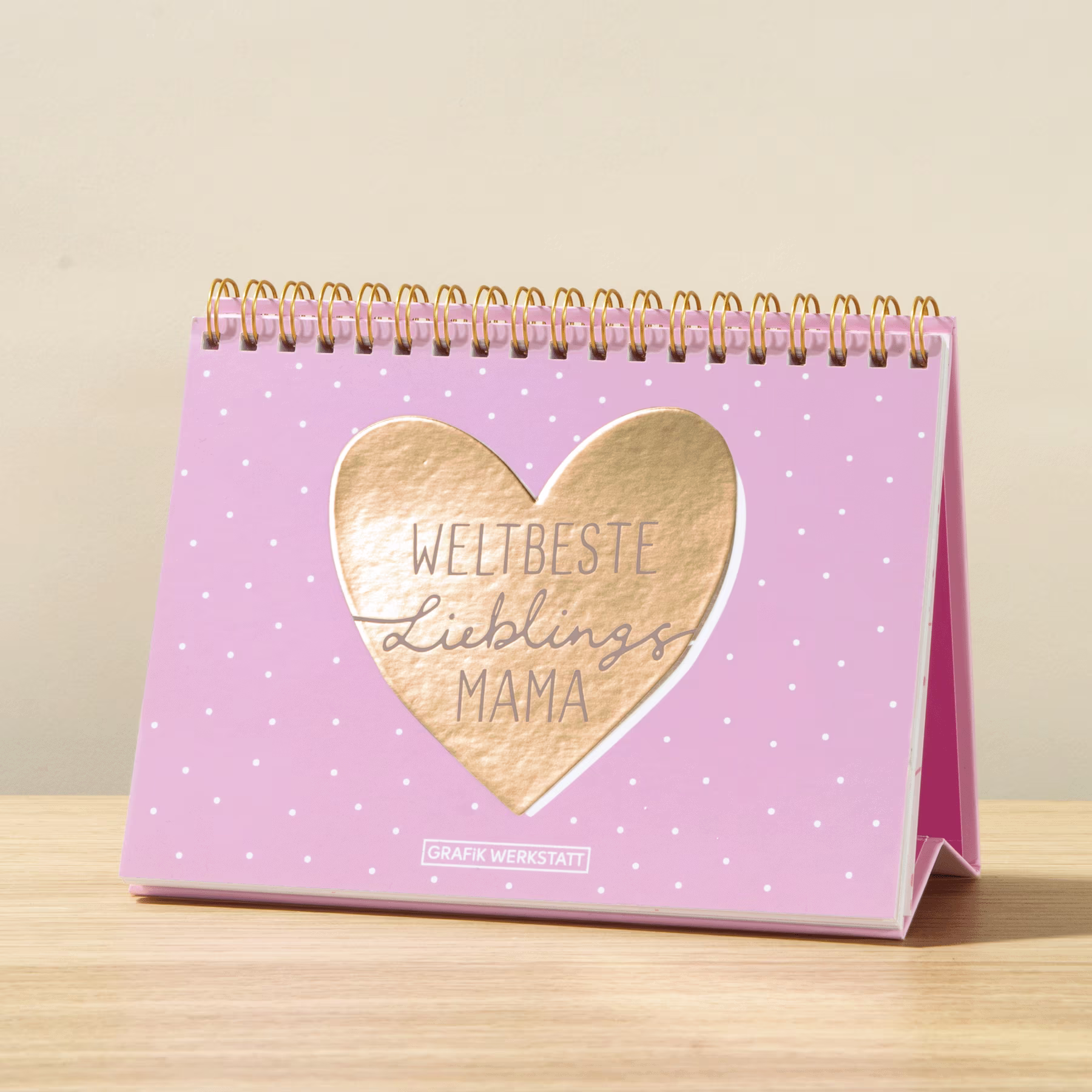 Ein rosa Spiralkalender mit einem herzförmigen Ausschnitt auf dem Cover, auf dem der deutsche Text „Weltbeste Lieblingsmama“ zu sehen ist. (Beschreibungstext KI-generiert)