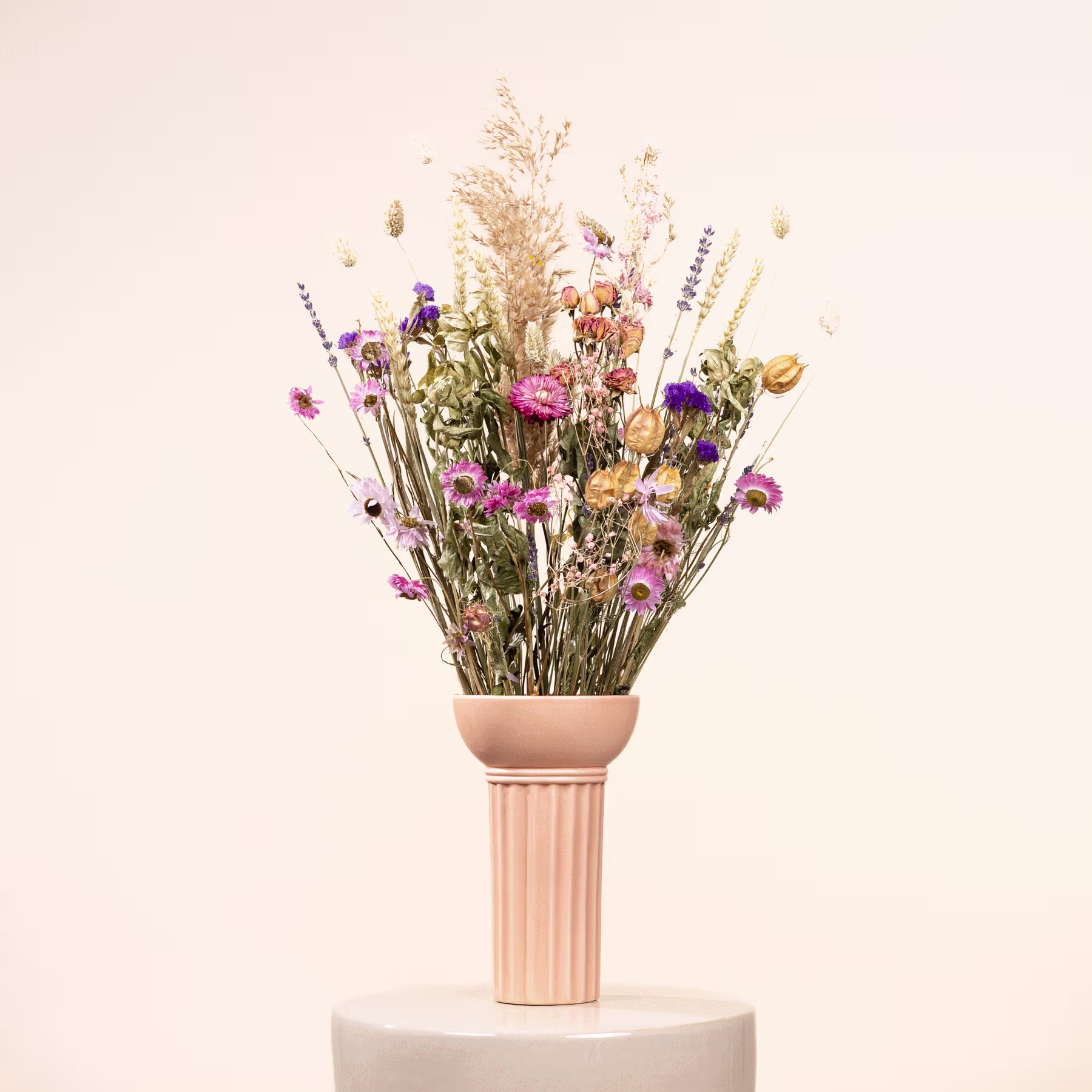 Ein Strauß leuchtender Trockenblumen und Gräser steht hoch in einer Keramikvase vor einem schlichten, hellen Hintergrund. (Beschreibungstext KI-generiert)
