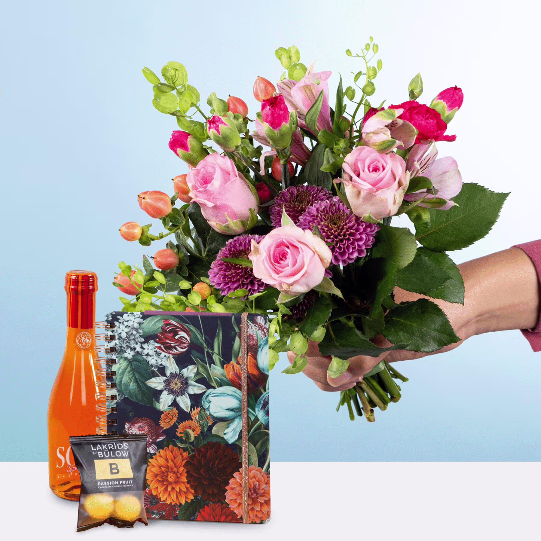Ein farbenfroher Strauß aus rosa Rosen, orangefarbenen Blumen und Grünpflanzen wird vor einer Geschenktüte mit Blumenmuster gehalten, begleitet von einer Flasche orangefarbener Flüssigkeit. (Beschreibungstext KI-generiert)