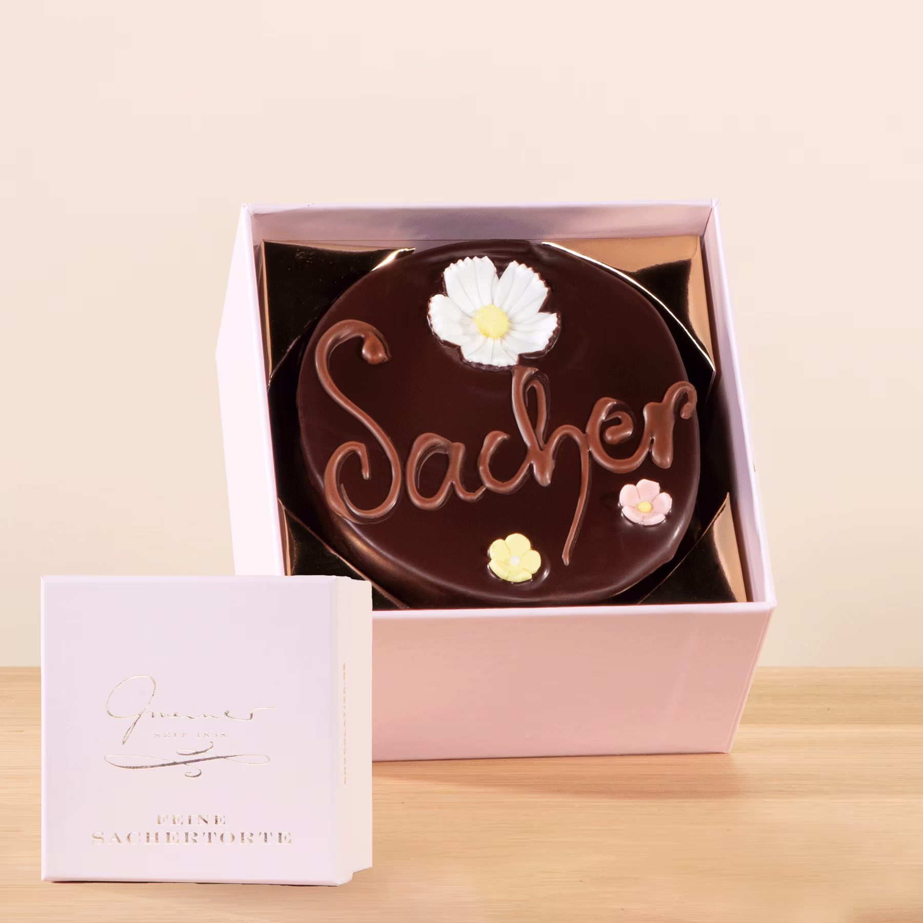 Eine Schokoladentorte mit der Aufschrift „Sacher“, dekoriert mit weißen Blumen, steht in einer rosa Schachtel auf einer Holzoberfläche. (Beschreibungstext KI-generiert)