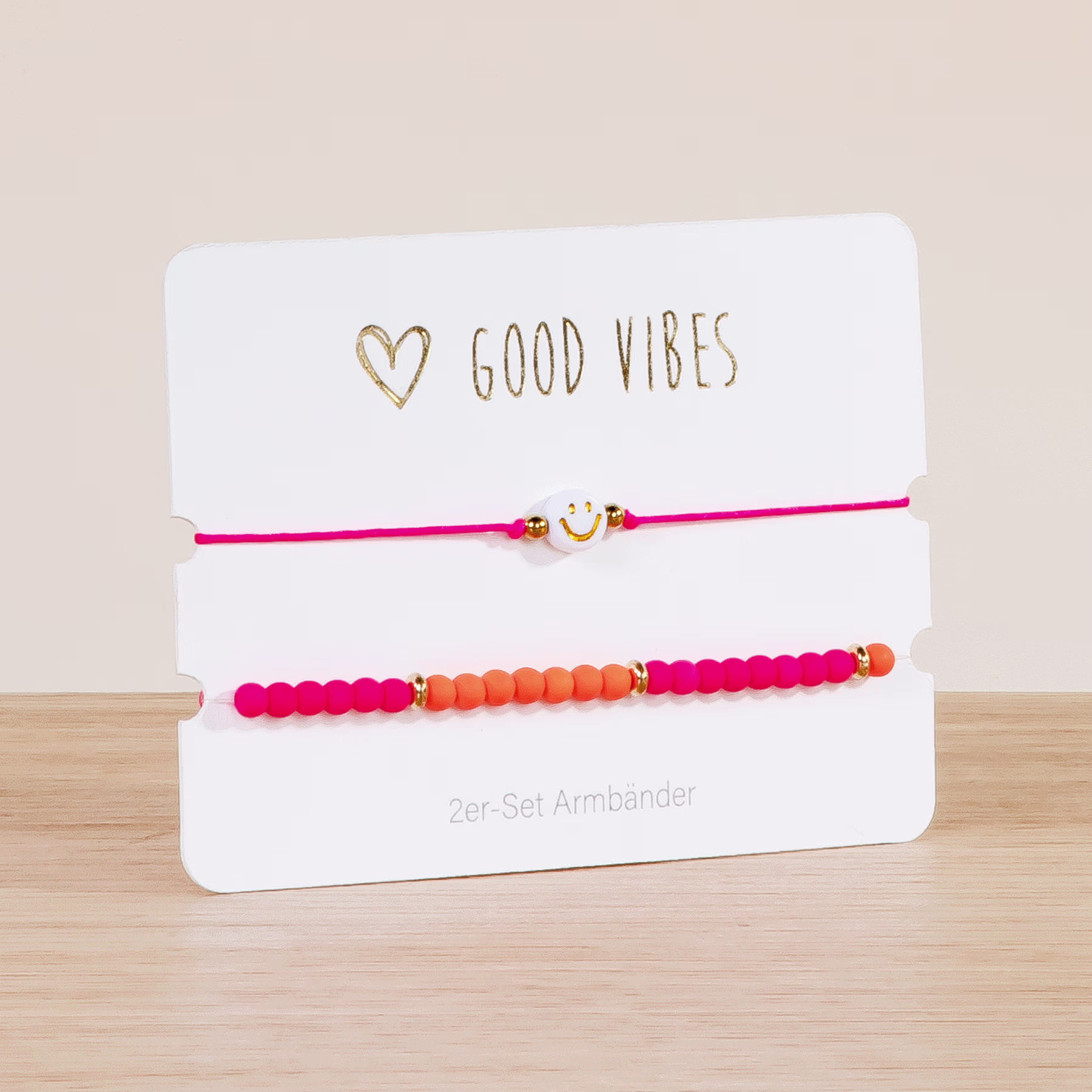 Das Bild zeigt ein Set bunter Perlenarmbänder auf einer weißen Karte mit dem goldenen Schriftzug „GOOD VIBES“. Die Armbänder sind optisch ansprechend angeordnet, wobei eine Mischung aus rosa, orangefarbenen und roten Perlen eine lebendige und fröhliche Ästhetik erzeugt. (Beschreibungstext KI-generiert)