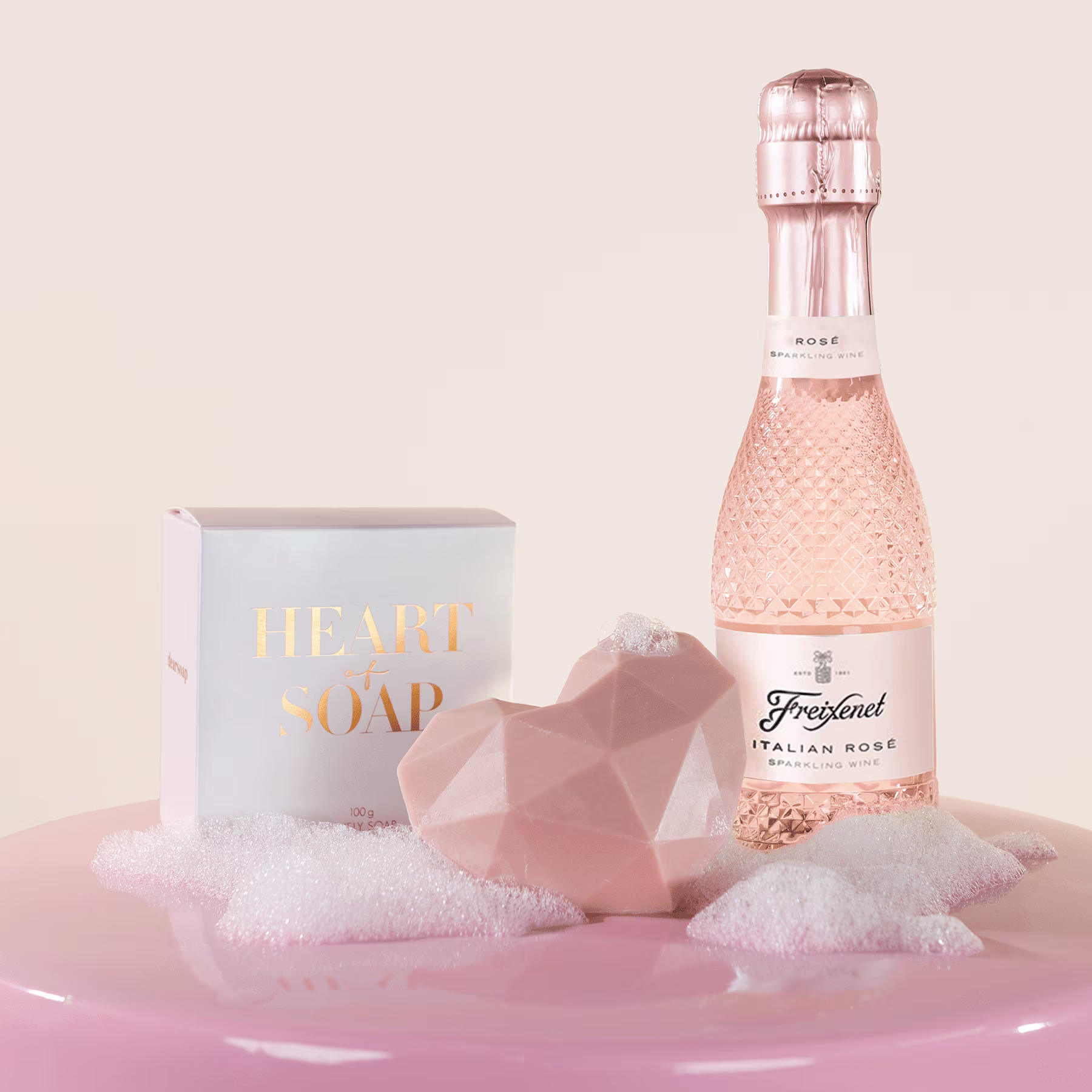 Eine rosa getönte Glasflasche Sekt, eine weiße Schachtel mit der Aufschrift „Luxusseife“ und ein rosafarbener, geometrisch geformter Gegenstand stehen auf einer weichen, flauschigen rosafarbenen Oberfläche. (Beschreibungstext KI-generiert)
