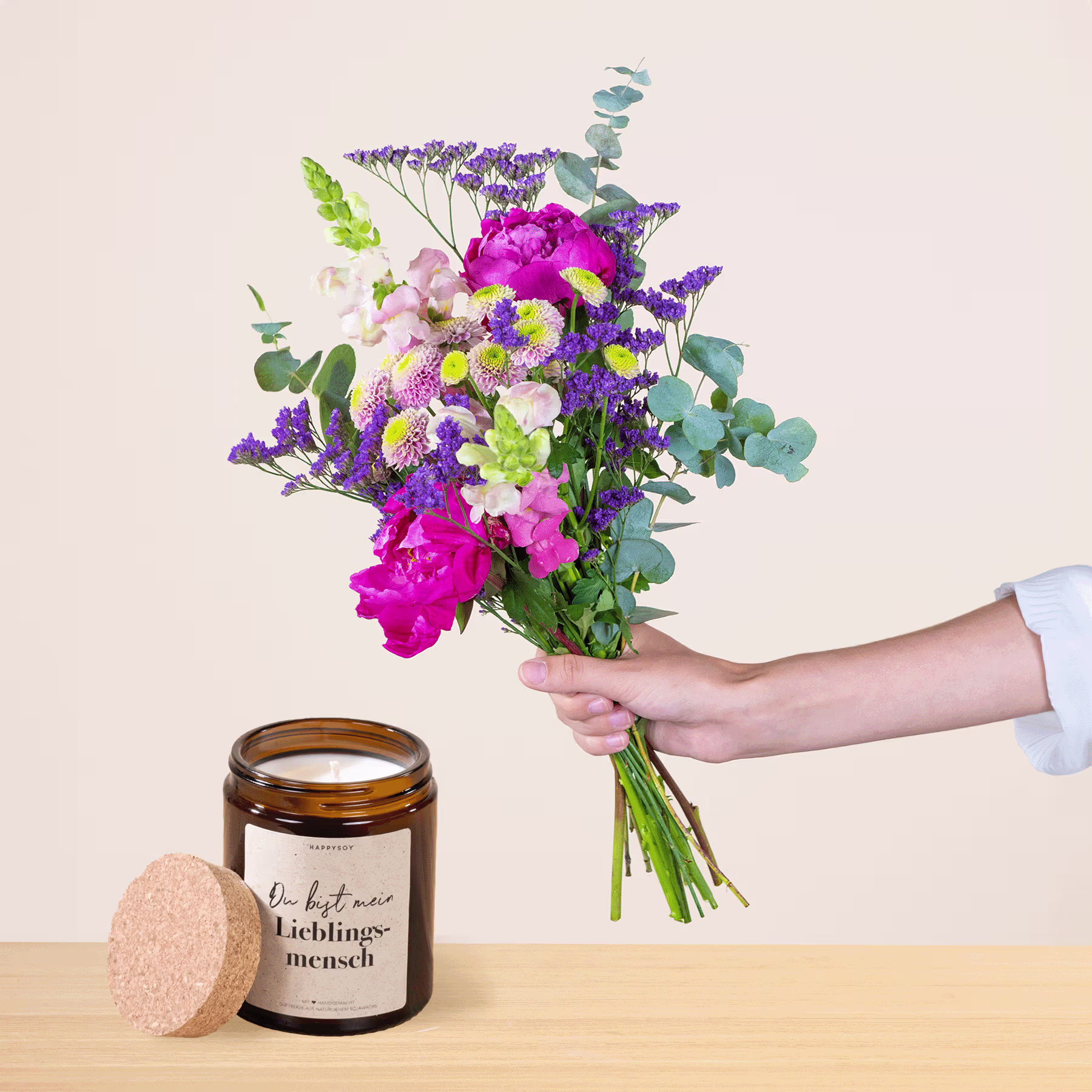 Eine Person hält einen leuchtenden Strauß bunter Blumen, darunter rosa Pfingstrosen, lila Lavendel und grüne Blätter, in der Hand, neben einer Kerze und einem herzförmigen Holzobjekt auf einem hellen Hintergrund. (Beschreibungstext KI-generiert)