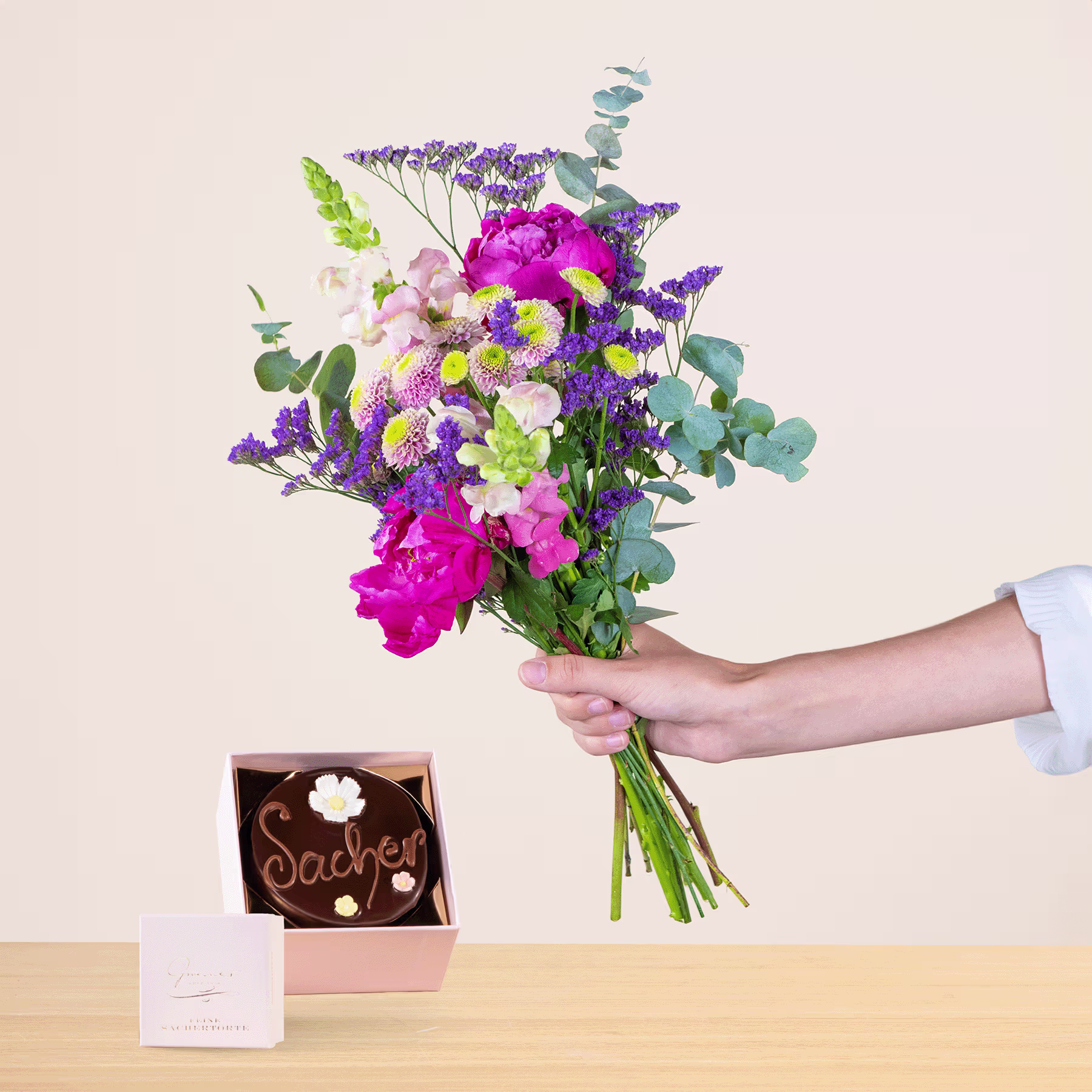 Eine Person hält einen leuchtend bunten Blumenstrauß, darunter rosa Pfingstrosen, lila Lavendel und grüne Blätter, in der Hand. Im Hintergrund liegen auf dem Tisch eine kleine Pralinenschachtel und eine Grußkarte. (Beschreibungstext KI-generiert)