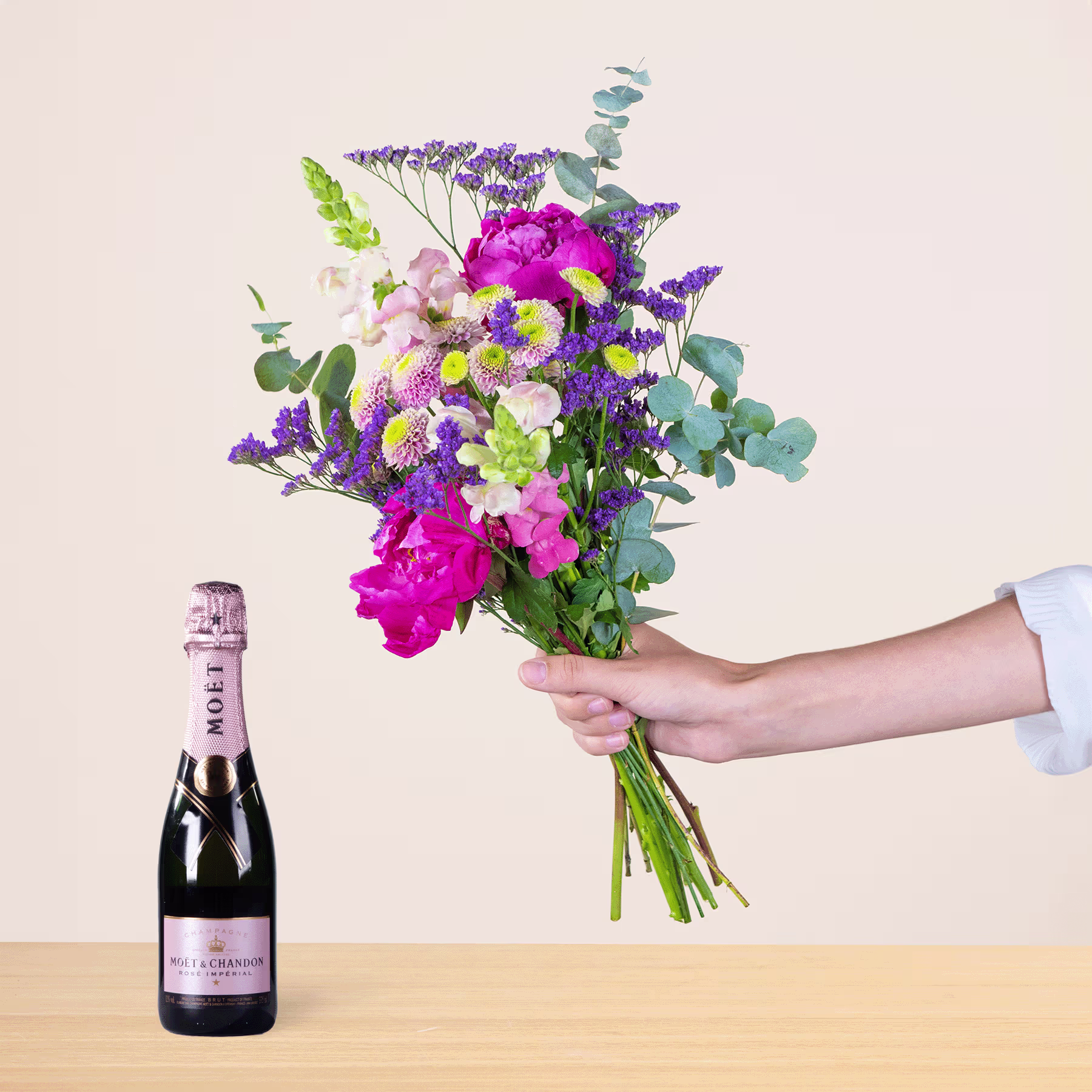 Ein leuchtender Strauß bunter Blumen, darunter rosa Pfingstrosen, lila Lavendel und grünes Laub, wird neben einer Flasche Champagner auf einer Holzoberfläche in der Hand einer Person gehalten. (Beschreibungstext KI-generiert)