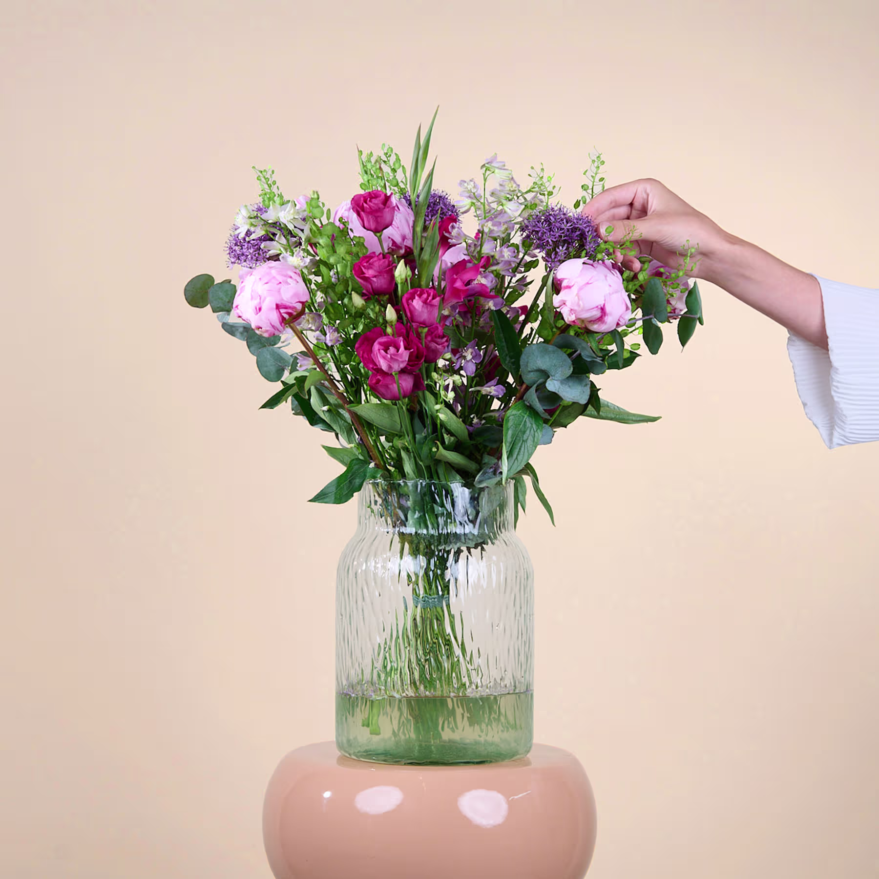 Ein farbenfroher Strauß rosa, violetter und weißer Blumen in einer Glasvase steht auf einer hellen Fläche vor einem schlichten Hintergrund. (Beschreibungstext KI-generiert)