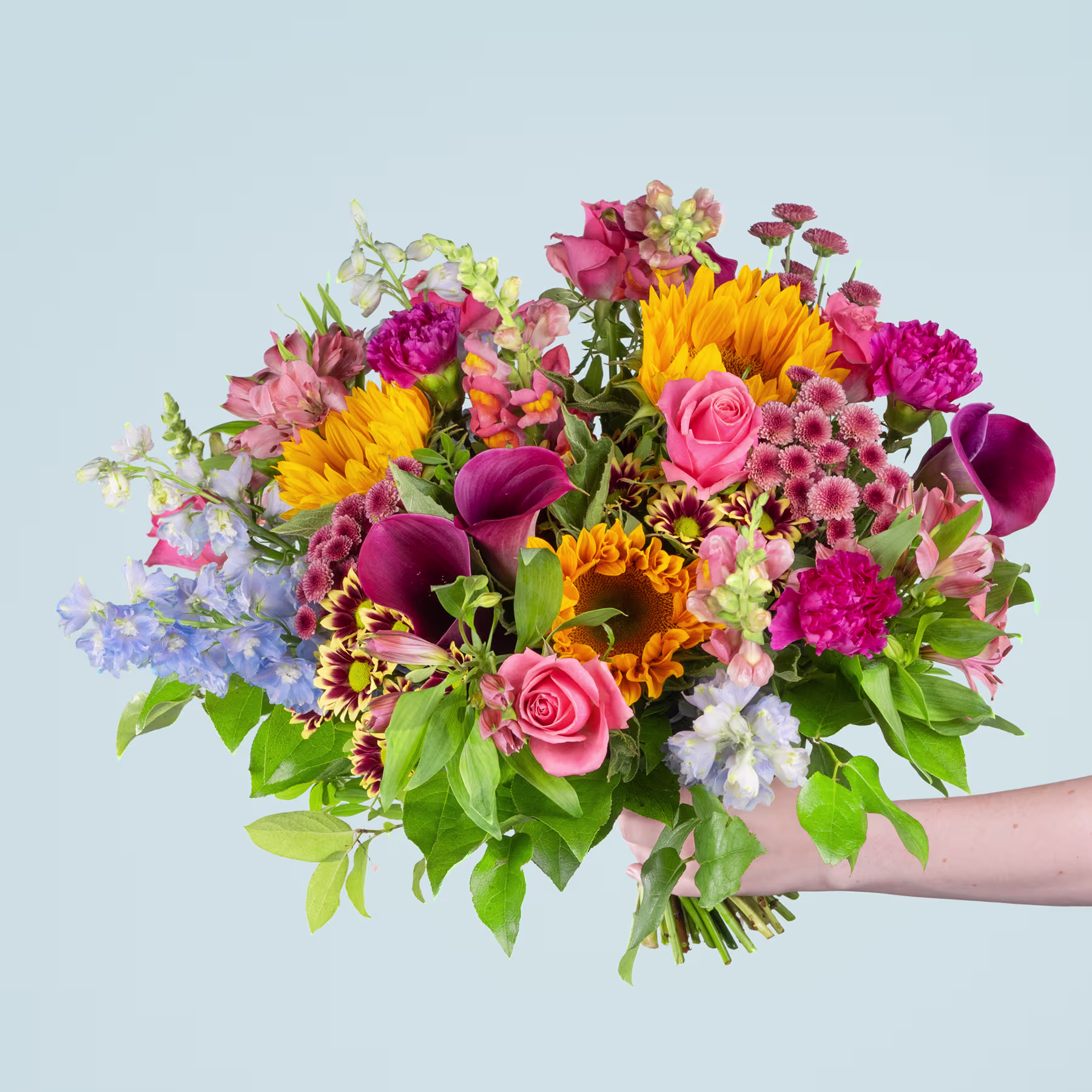 Ein lebendiger Strauß bunter Blumen, darunter Sonnenblumen, Rosen, Nelken und andere Blüten, in einer Hand gehalten vor einem schlichten hellblauen Hintergrund. (Beschreibungstext KI-generiert)