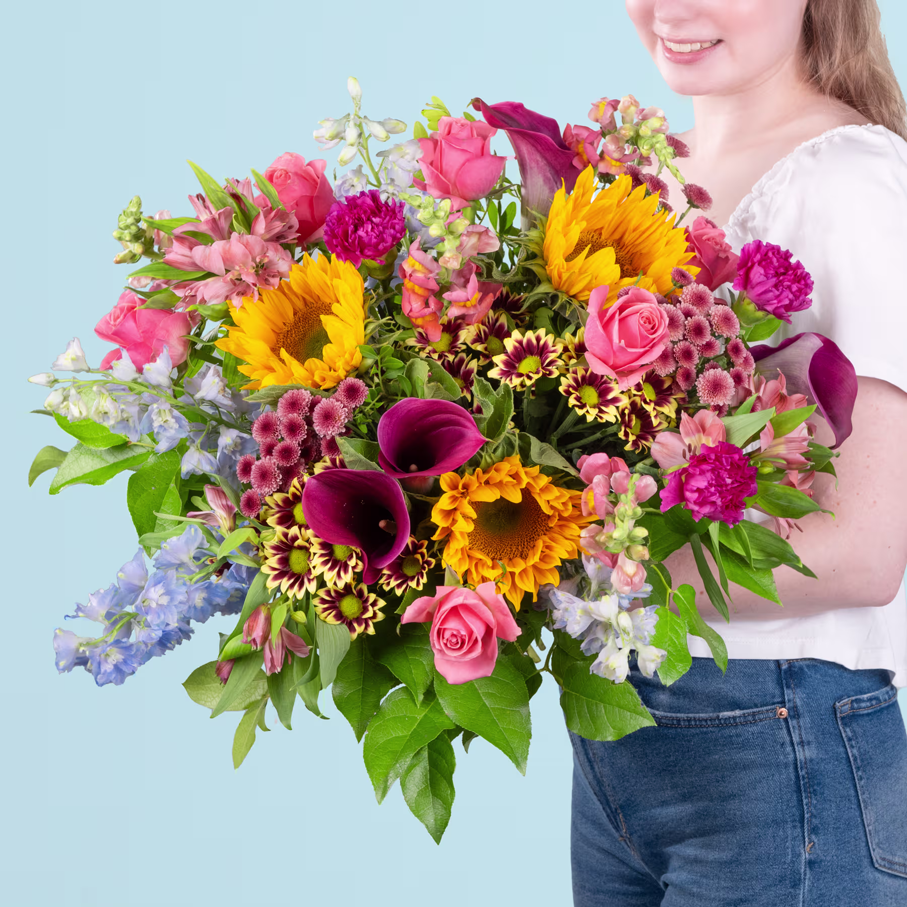 Ein leuchtender Strauß bunter Blumen, darunter Sonnenblumen, Rosen und andere Blüten, wird von einer Person in Jeans vor einem hellblauen Hintergrund gehalten. (Beschreibungstext KI-generiert)
