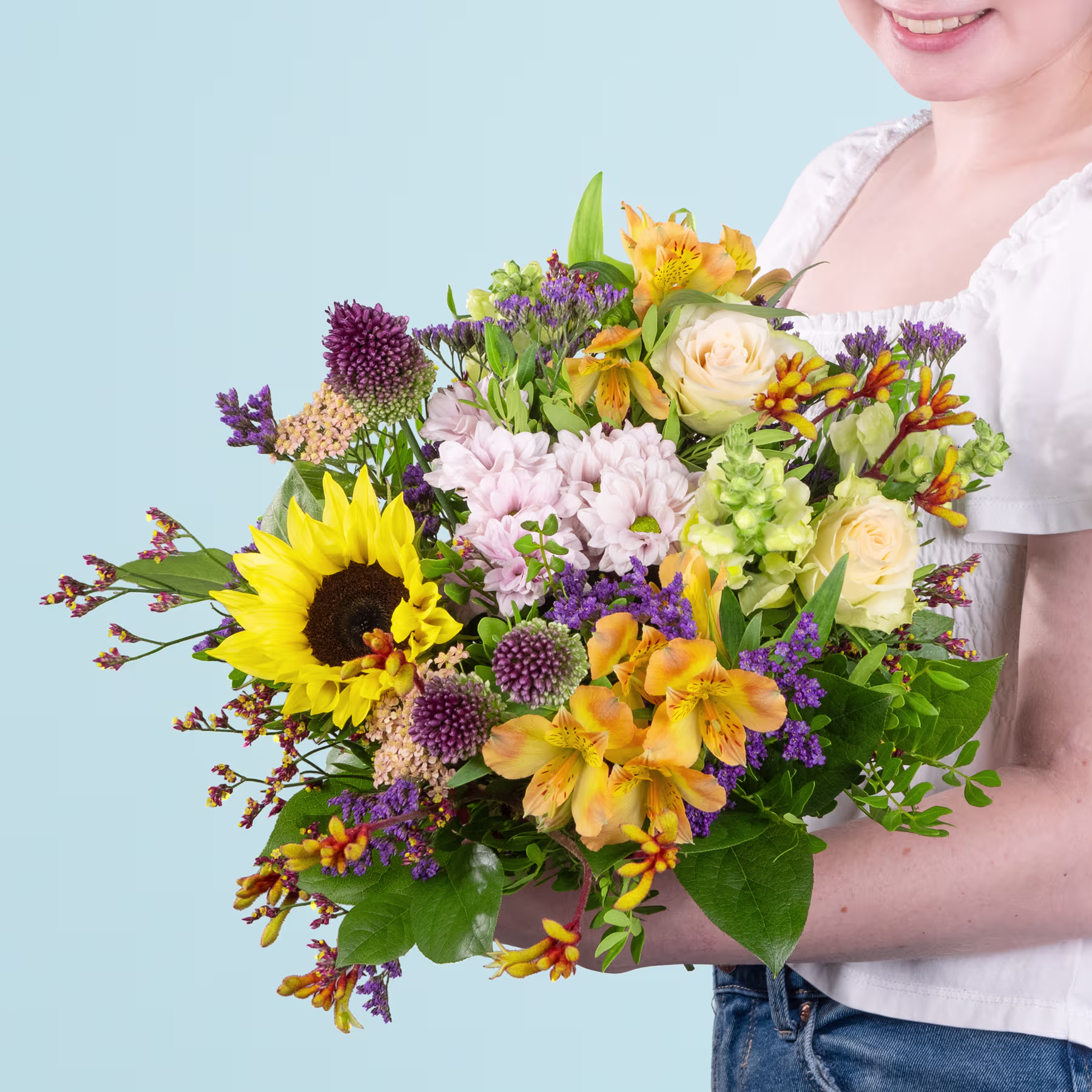 Ein leuchtender Strauß bunter Blumen, darunter Sonnenblumen, Rosen und verschiedene andere Blüten, wird von einer Person im Vordergrund vor einem hellblauen Hintergrund gehalten. (Beschreibungstext KI-generiert)