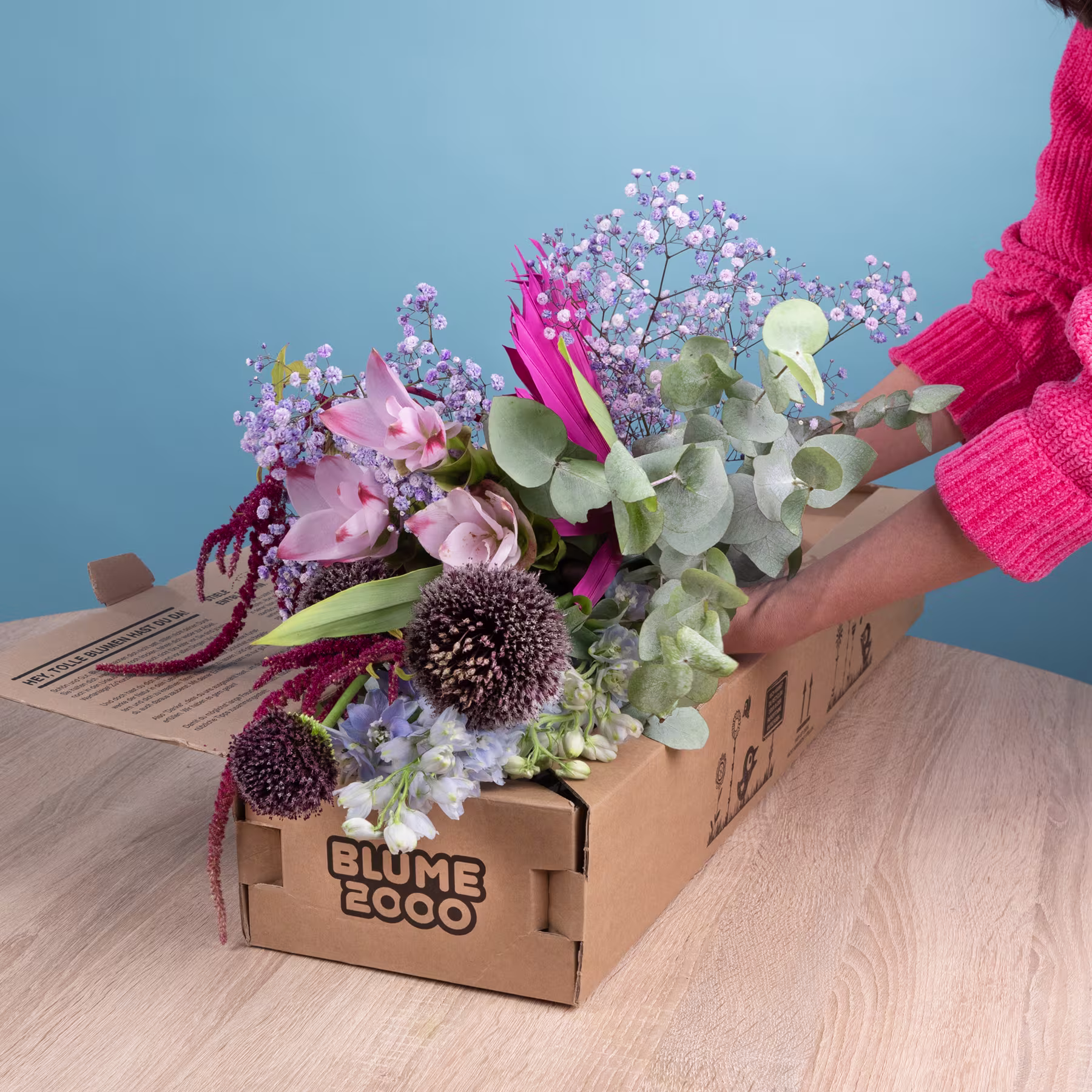 Eine Holzkiste mit einer farbenfrohen Auswahl an bunten Blumen, darunter rosa und weiße Blüten, steht auf einer Holzoberfläche vor einem blauen Hintergrund. (Beschreibungstext KI-generiert)