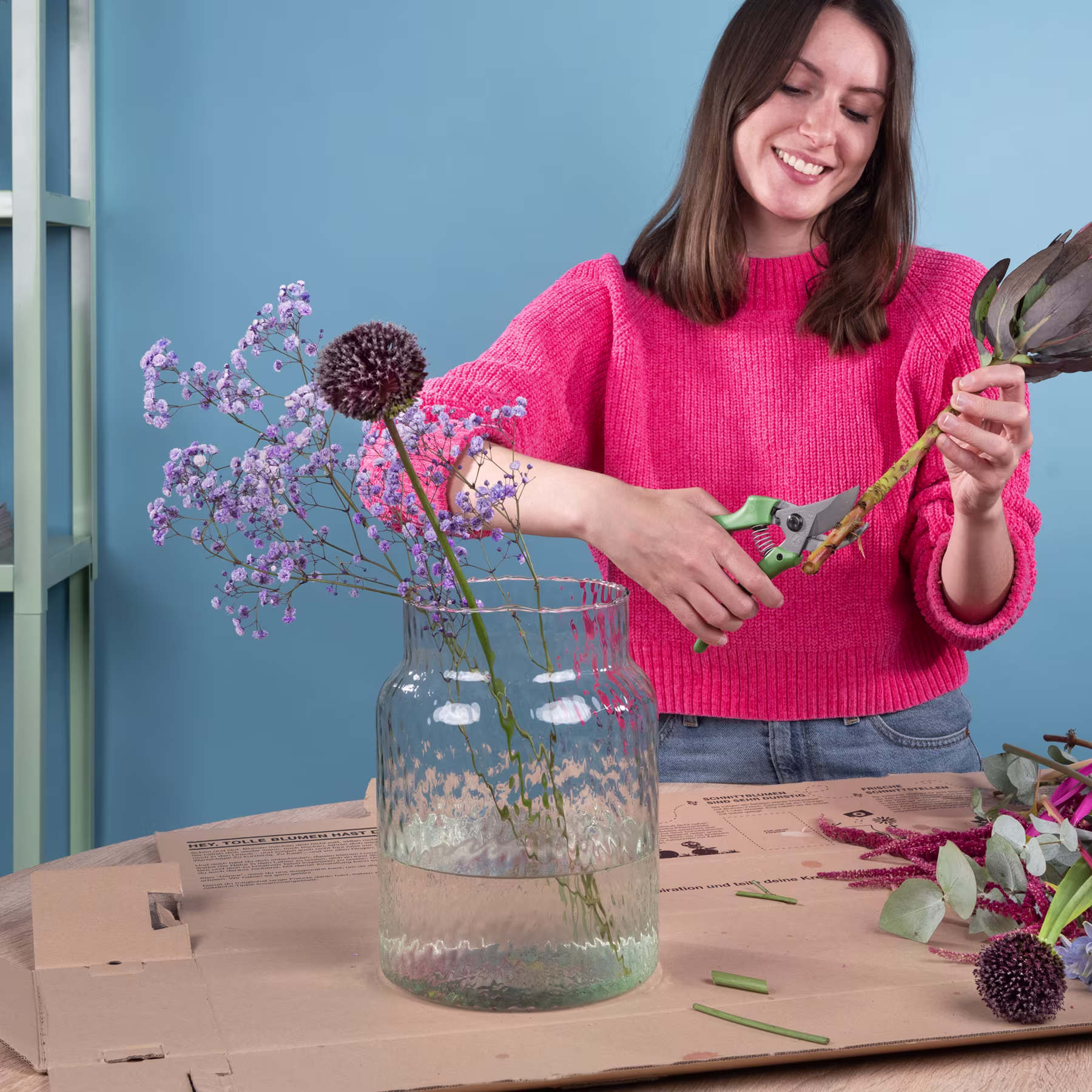 Eine junge Frau mit langen braunen Haaren lächelt, während sie einen Strauß lila Blumen in einer Glasvase auf einem Holztisch arrangiert, im Hintergrund eine blaue Wand. (Beschreibungstext KI-generiert)