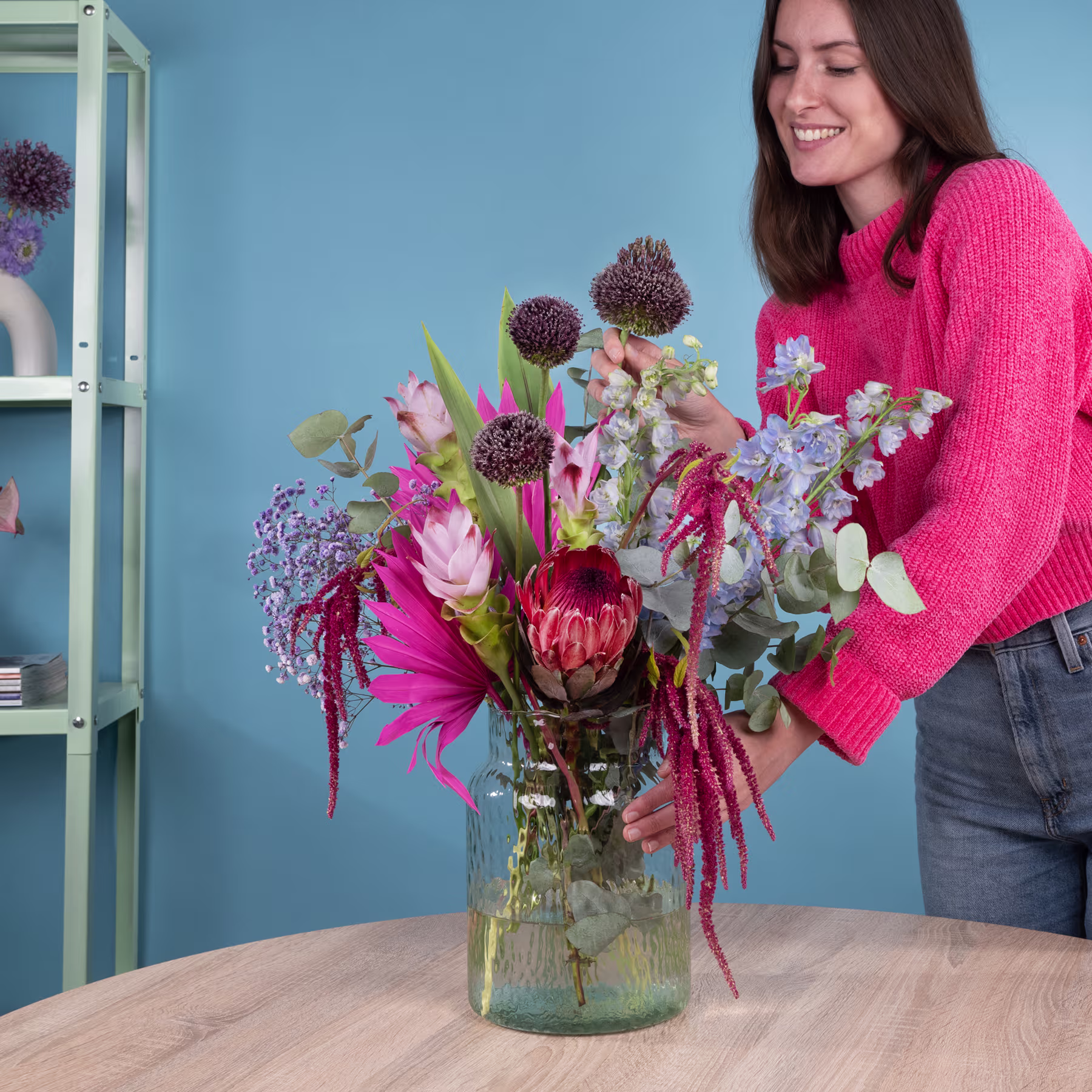 Eine junge Frau in einem leuchtend rosa Pullover steht vor einem blauen Hintergrund neben einer großen Vase mit einem leuchtend bunten Blumenstrauß, darunter Proteas, Rosen und andere Blüten. (Beschreibungstext KI-generiert)