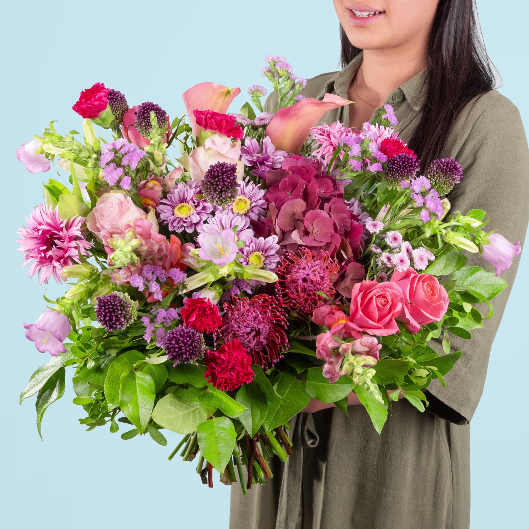 Ein leuchtender Strauß bunter Blumen, darunter Rosen, Nelken und Chrysanthemen, wird von einer lächelnden Frau in einer grünen Jacke vor einem schlichten blauen Hintergrund gehalten. (Beschreibungstext KI-generiert)