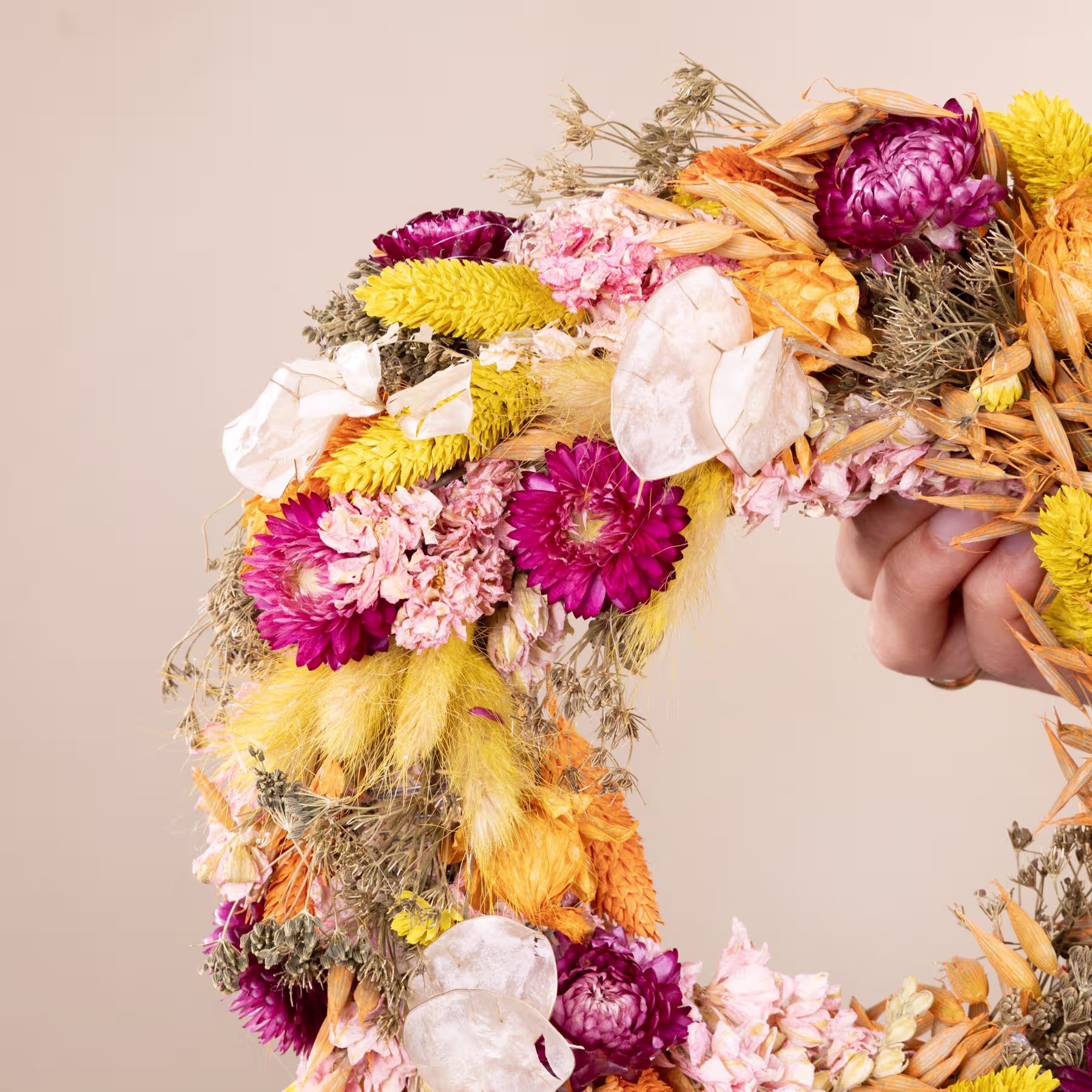 Ein lebendiger und farbenfroher Blumenkranz aus verschiedenen Blumen, darunter rosa und gelbe Chrysanthemen, weiße Pfingstrosen und getrocknetes Laub, wird im Vordergrund vor einem neutralen beigen Hintergrund gehalten. (Beschreibungstext KI-generiert)