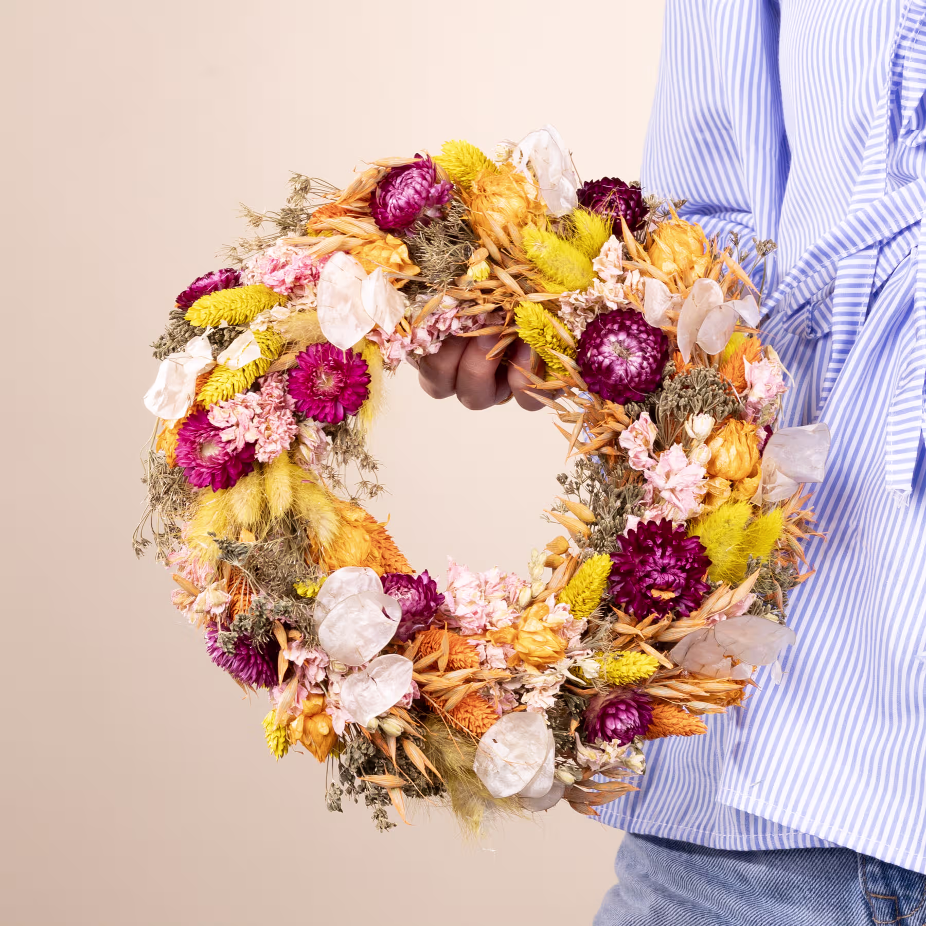 Ein farbenfroher Herbstkranz aus getrockneten Blumen und Blättern, gehalten von einer Person in einem gestreiften Hemd vor einem neutralen Hintergrund. (Beschreibungstext KI-generiert)