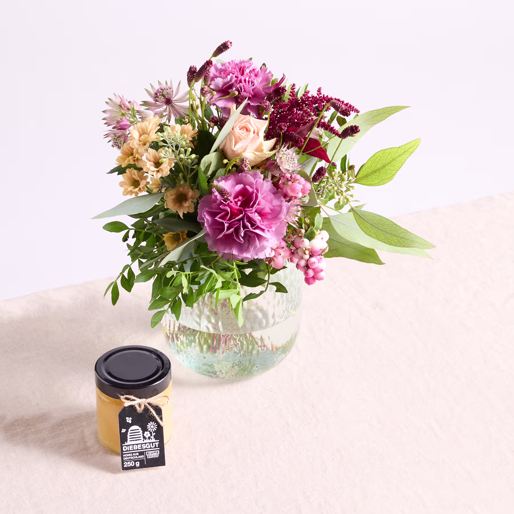 Ein leuchtender Strauß bunter Blumen, darunter rosa Pfingstrosen, gelbe Gänseblümchen und violette Chrysanthemen, wird in einer Glasvase auf einer hellen Oberfläche präsentiert, im Vordergrund steht ein kleines schwarzes Gefäß. (Beschreibungstext KI-generiert)