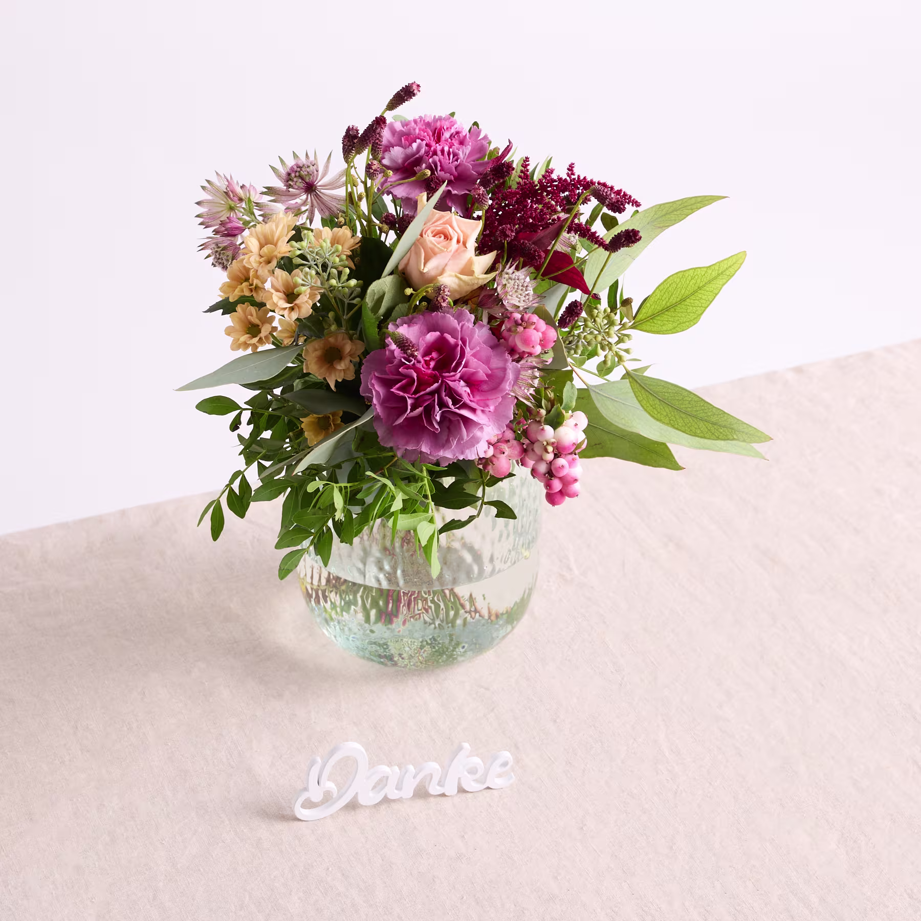 Ein farbenfroher Strauß verschiedener Blumen, darunter Pfingstrosen, Gänseblümchen und andere Blüten, wird in einer Glasvase auf einer hellen Oberfläche präsentiert. (Beschreibungstext KI-generiert)