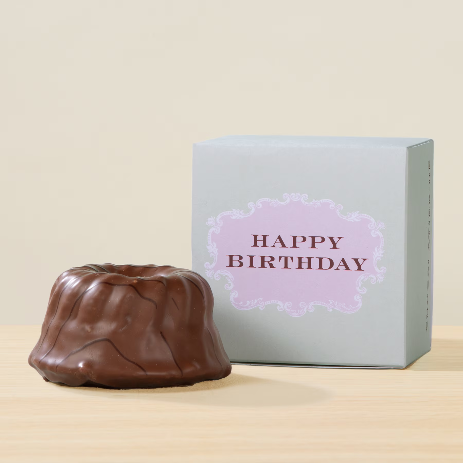 Auf einer Holzoberfläche steht ein Dessert in Form eines Schokoladenkuchens, begleitet von einer weißen Geschenkbox mit dem Aufdruck „HAPPY BIRTHDAY“. (Beschreibungstext KI-generiert)